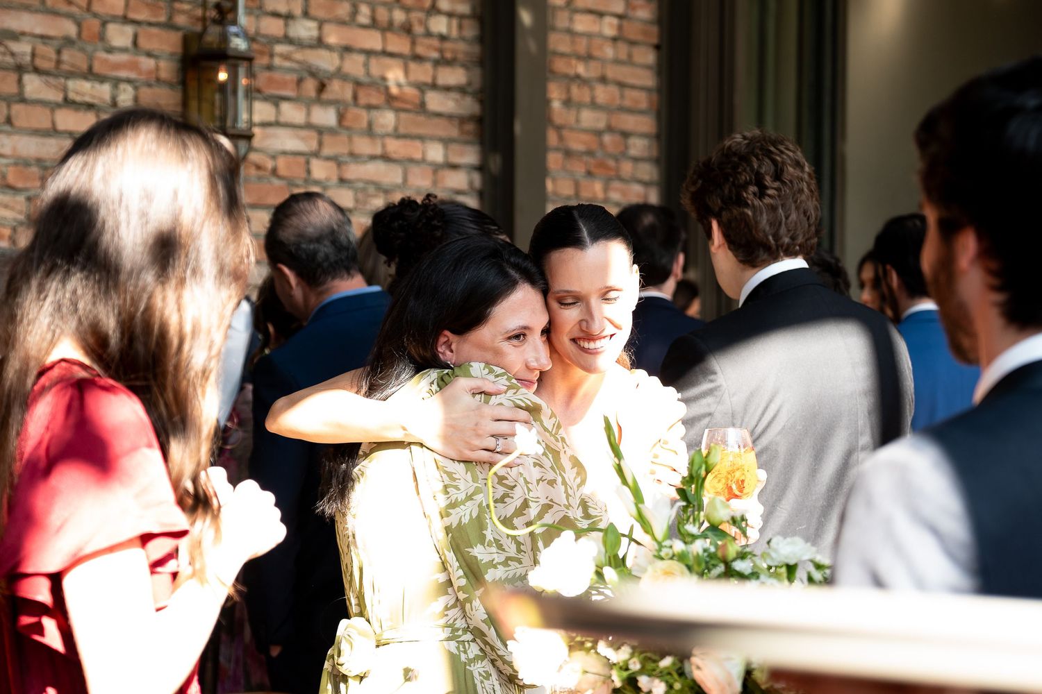 Bride holding yellow bouquet receives warm hug from guest at outdoor wedding reception near brick building.