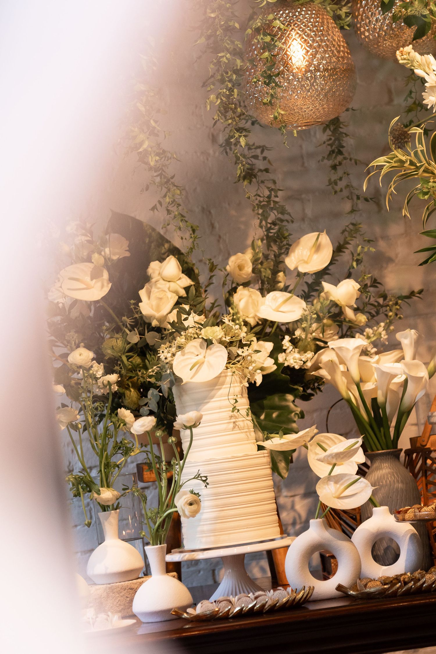 Elegant wedding cake display surrounded by white flowers, greenery, and wicker pendant lights on a decorated table.