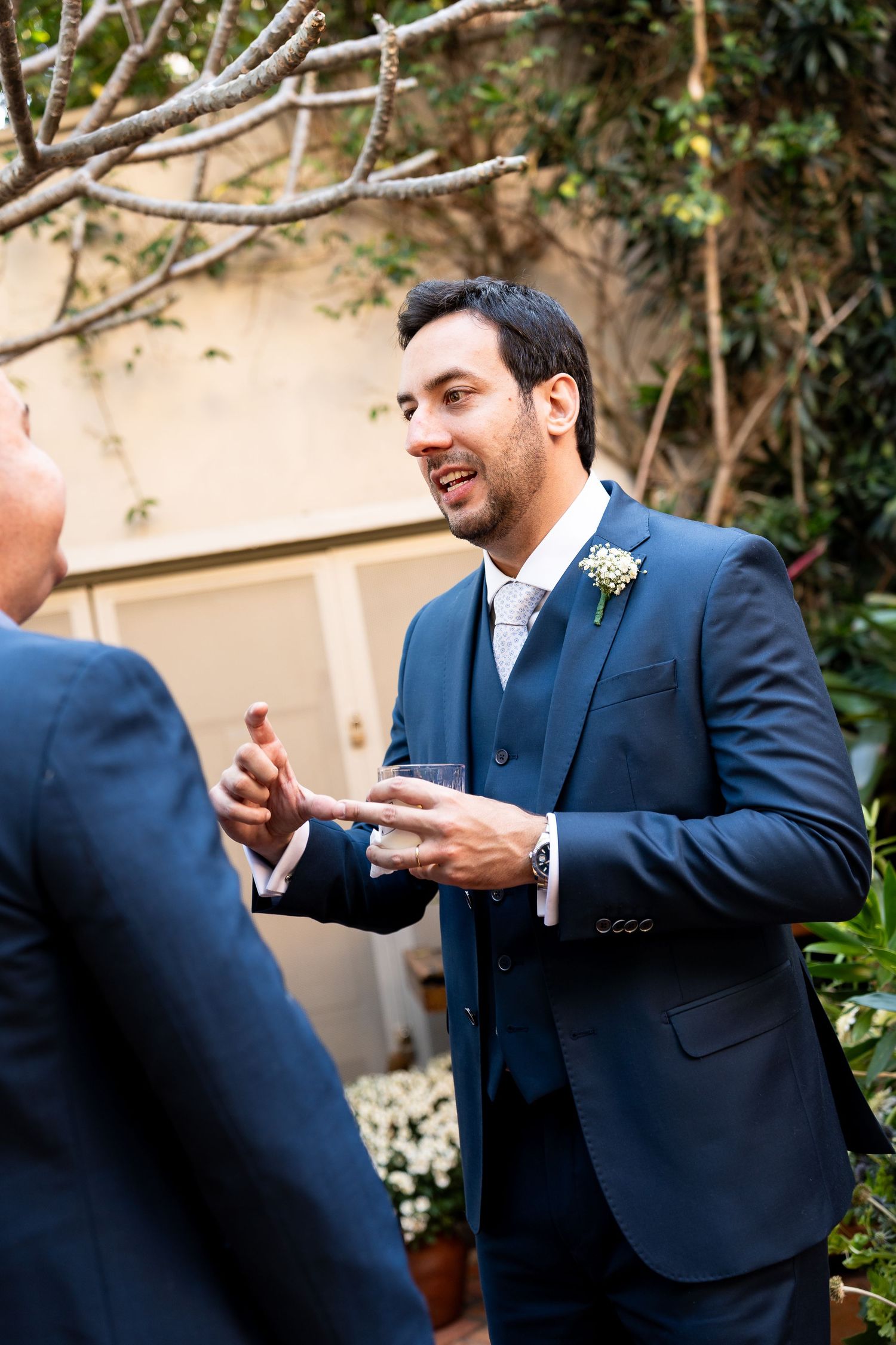 Groom in navy blue three-piece suit with boutonniere holds drink while conversing outdoors at wedding reception.
