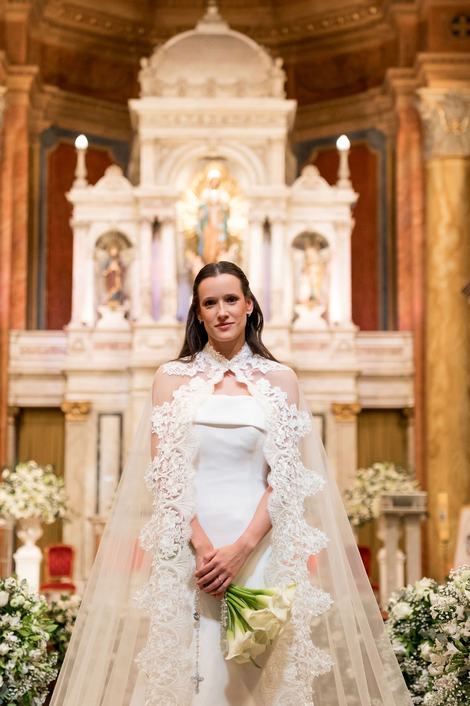 Elegant bride in white gown with lace cape holding bouquet, standing before ornate church altar.
