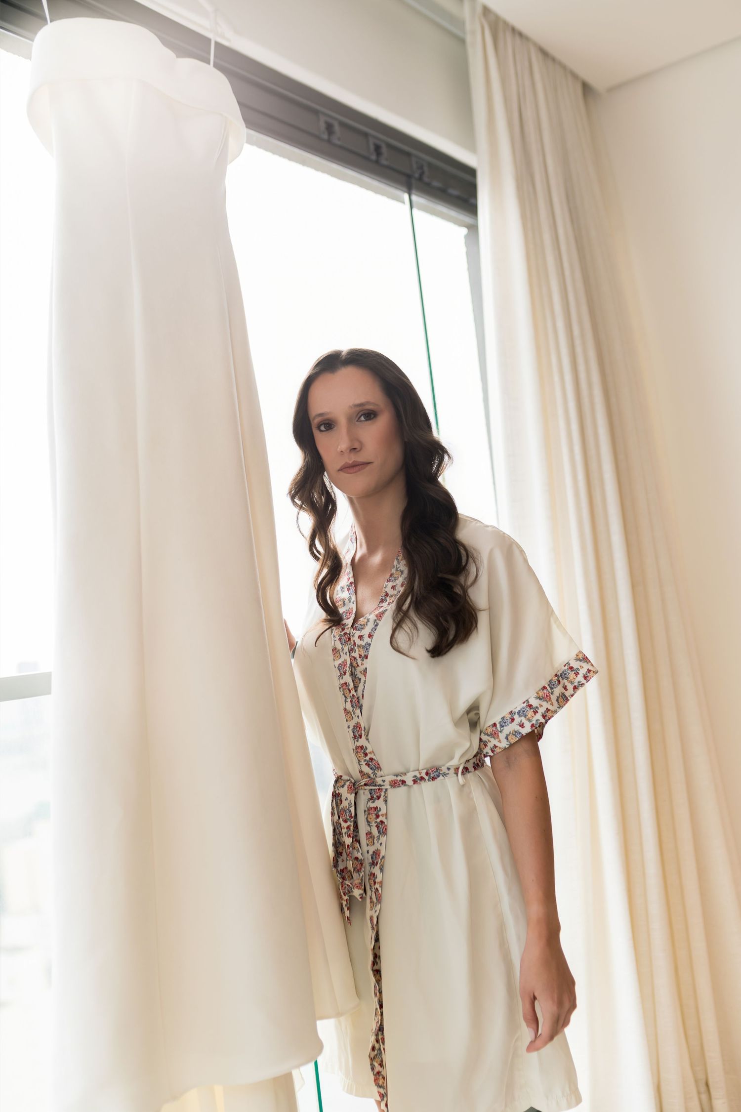 Bride in white robe gazing at wedding dress hanging by window with sheer curtains in bridal suite.