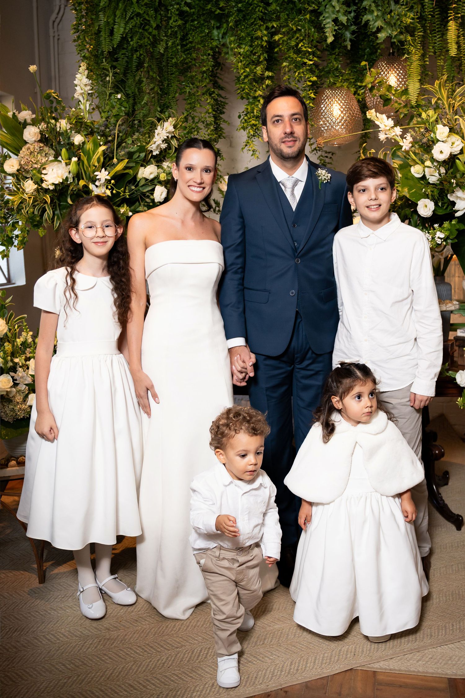 Bride and groom pose with four children in white attire at an elegant wedding ceremony with floral decor.