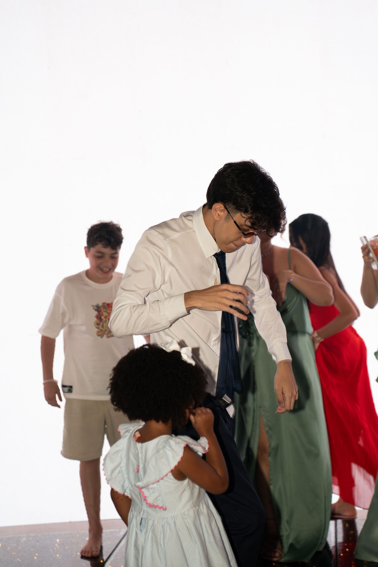 Man in white shirt and tie bending down to interact with a young girl in a white dress at a formal event.