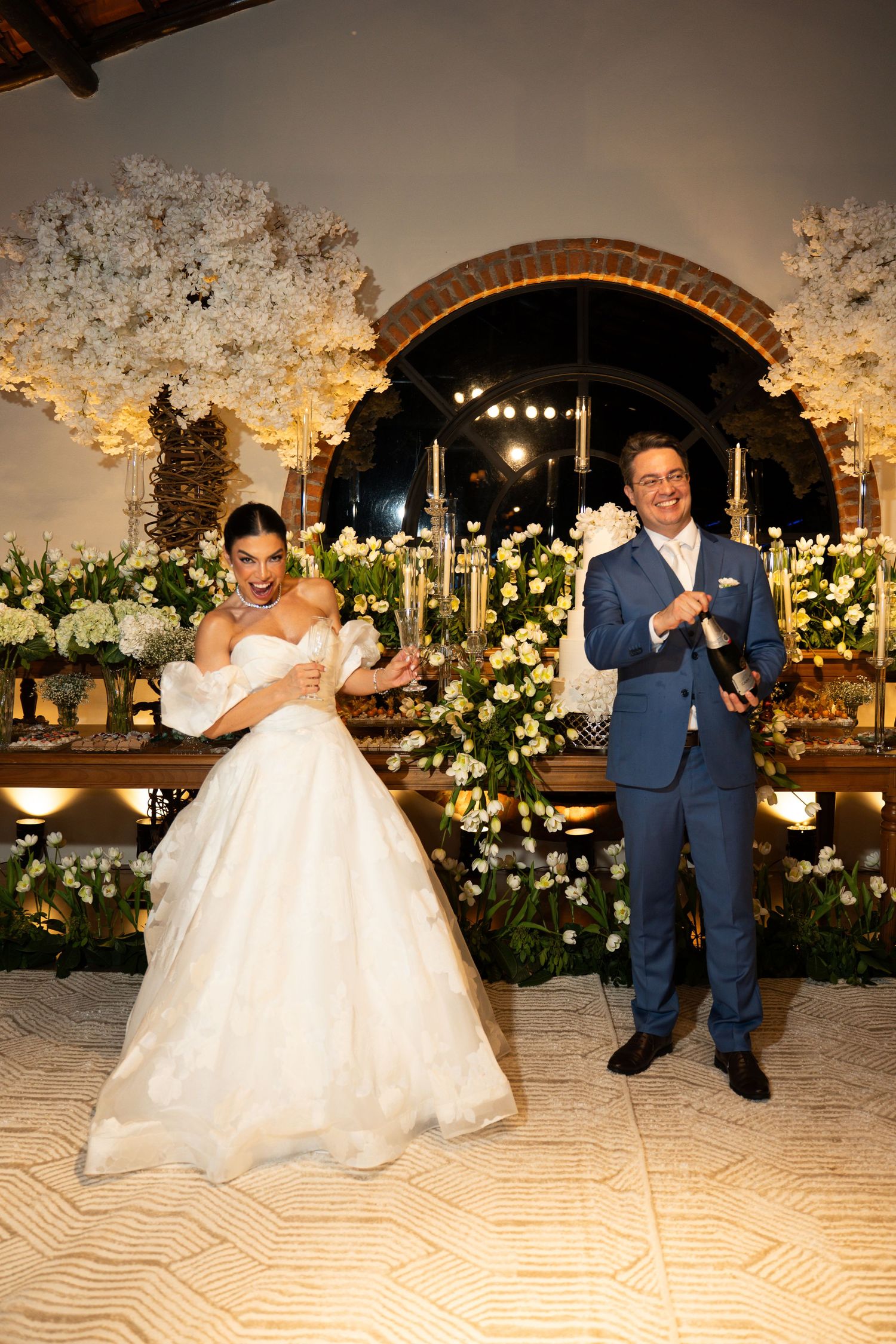 Bride in white ball gown and groom in blue suit celebrate joyfully at elegant floral wedding reception.