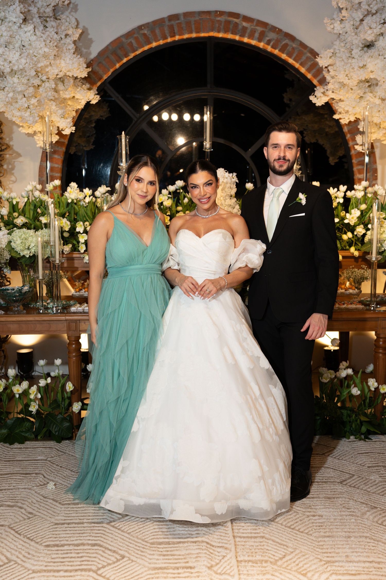 Bride in white ball gown poses with groom in black tuxedo and bridesmaid in teal dress at elegant floral wedding reception.
