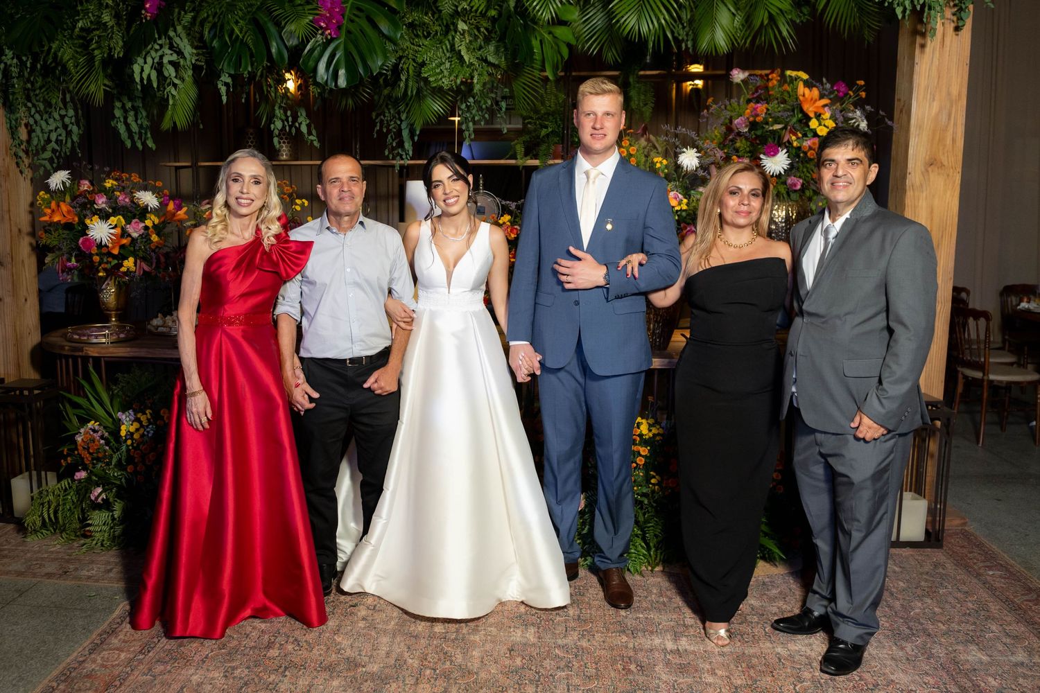 A wedding party of six poses together, with the bride in white and guests in formal attire amid lush tropical floral decor.