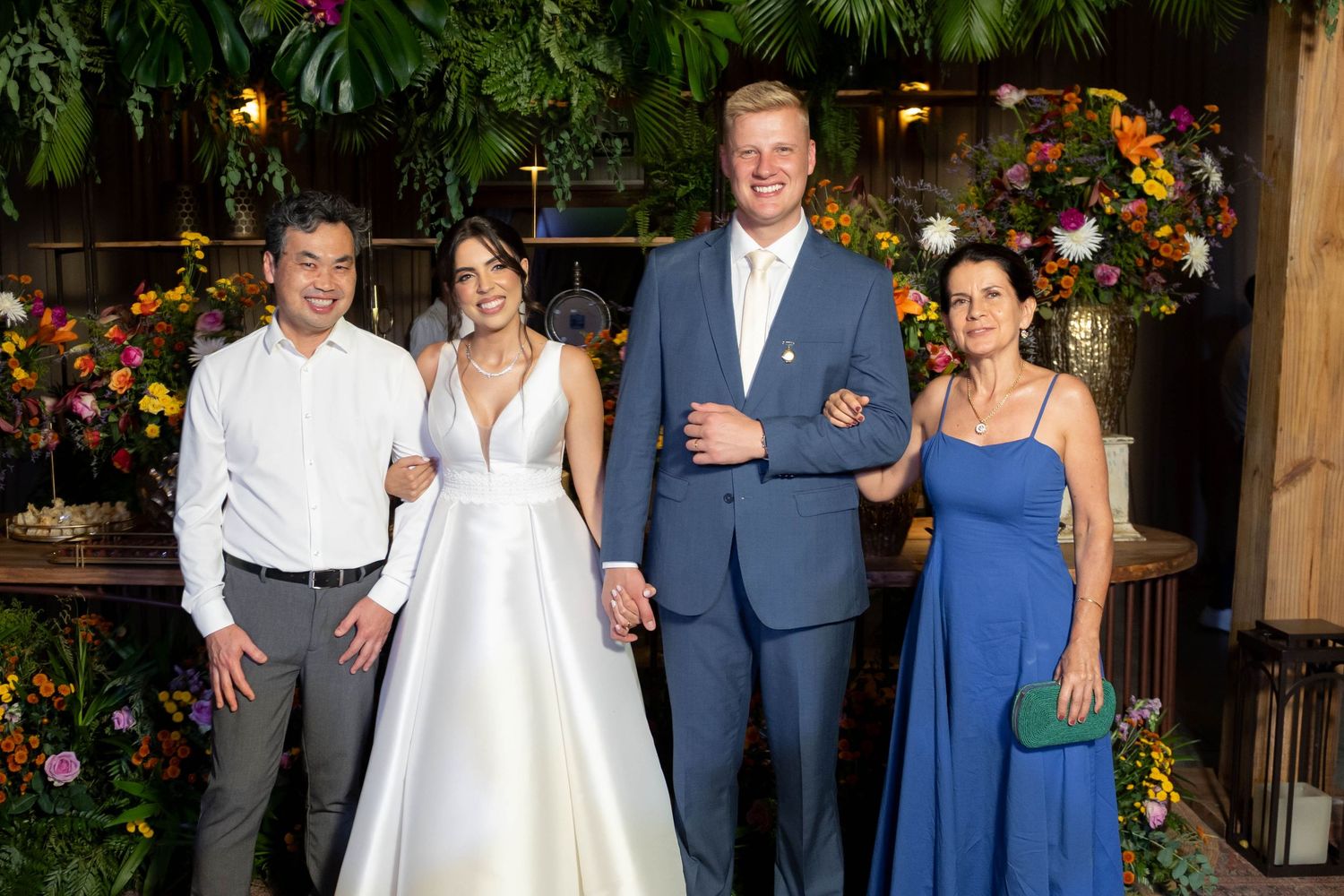 Wedding party of four posing indoors with floral decor, bride in white gown, groom in blue suit, guests in formal attire.