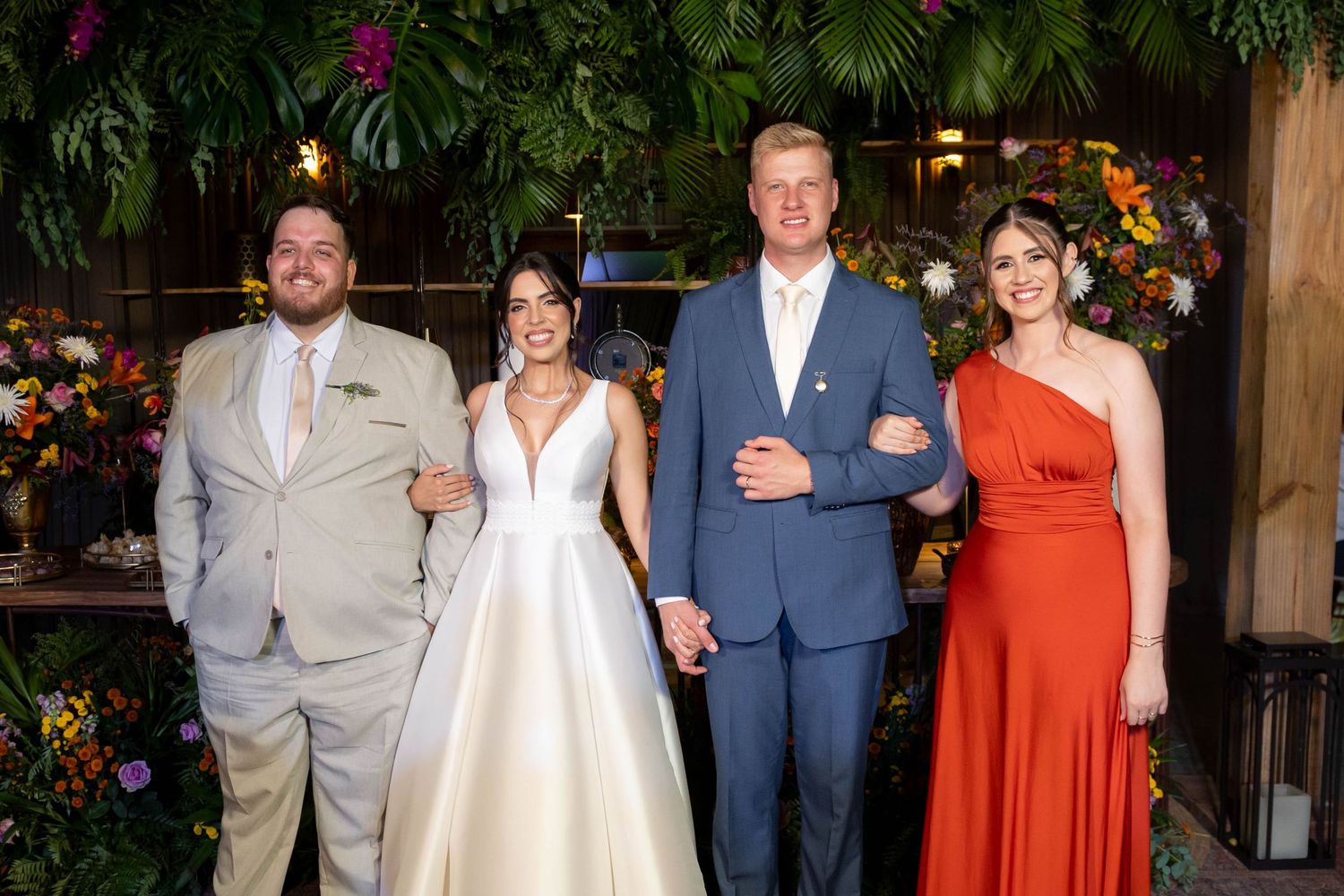 Wedding party of four posing indoors with lush greenery backdrop, bride in white gown flanked by guests in formal attire.