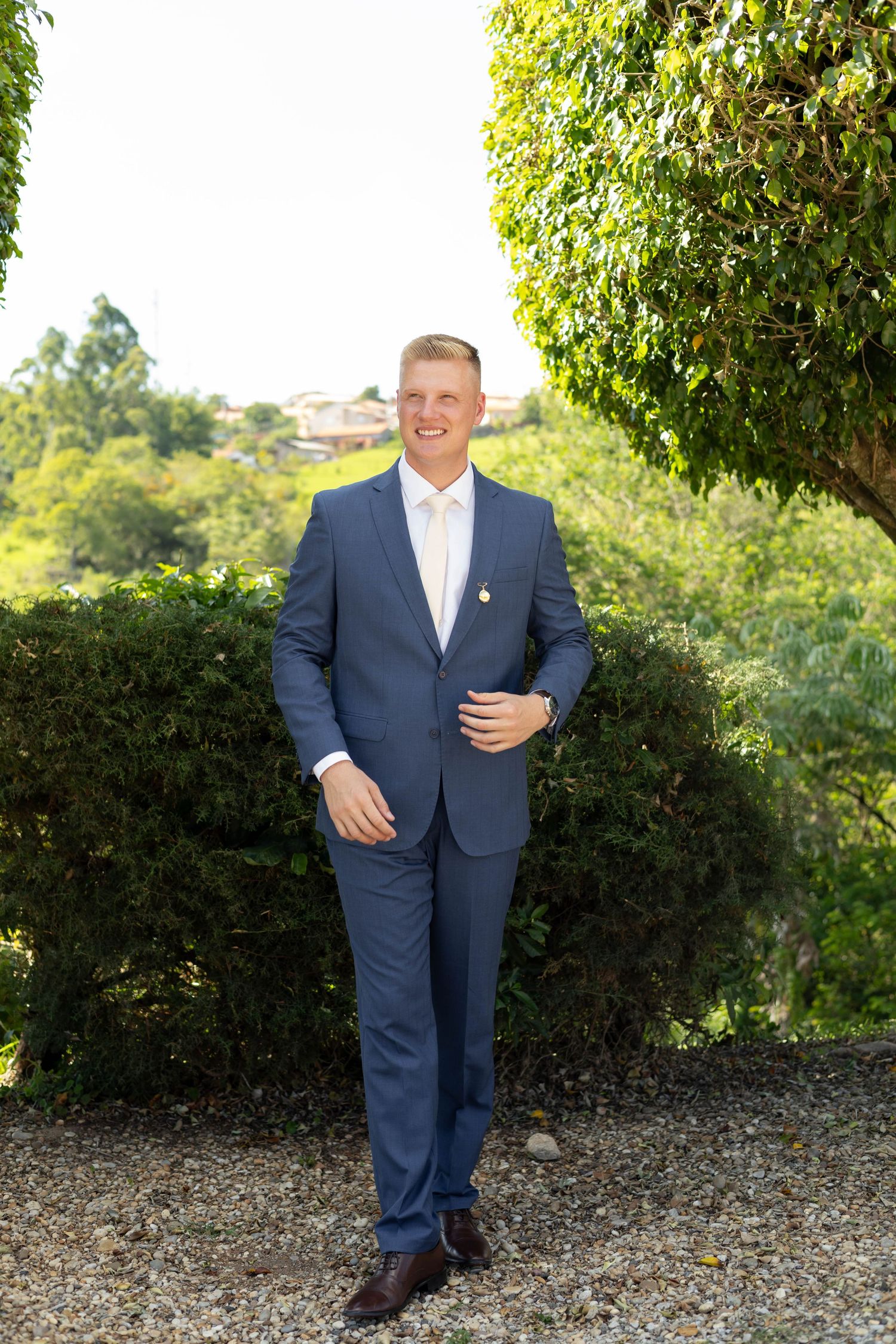 Smiling man in a blue suit and white tie standing outdoors among green hedges and trees on a sunny day.