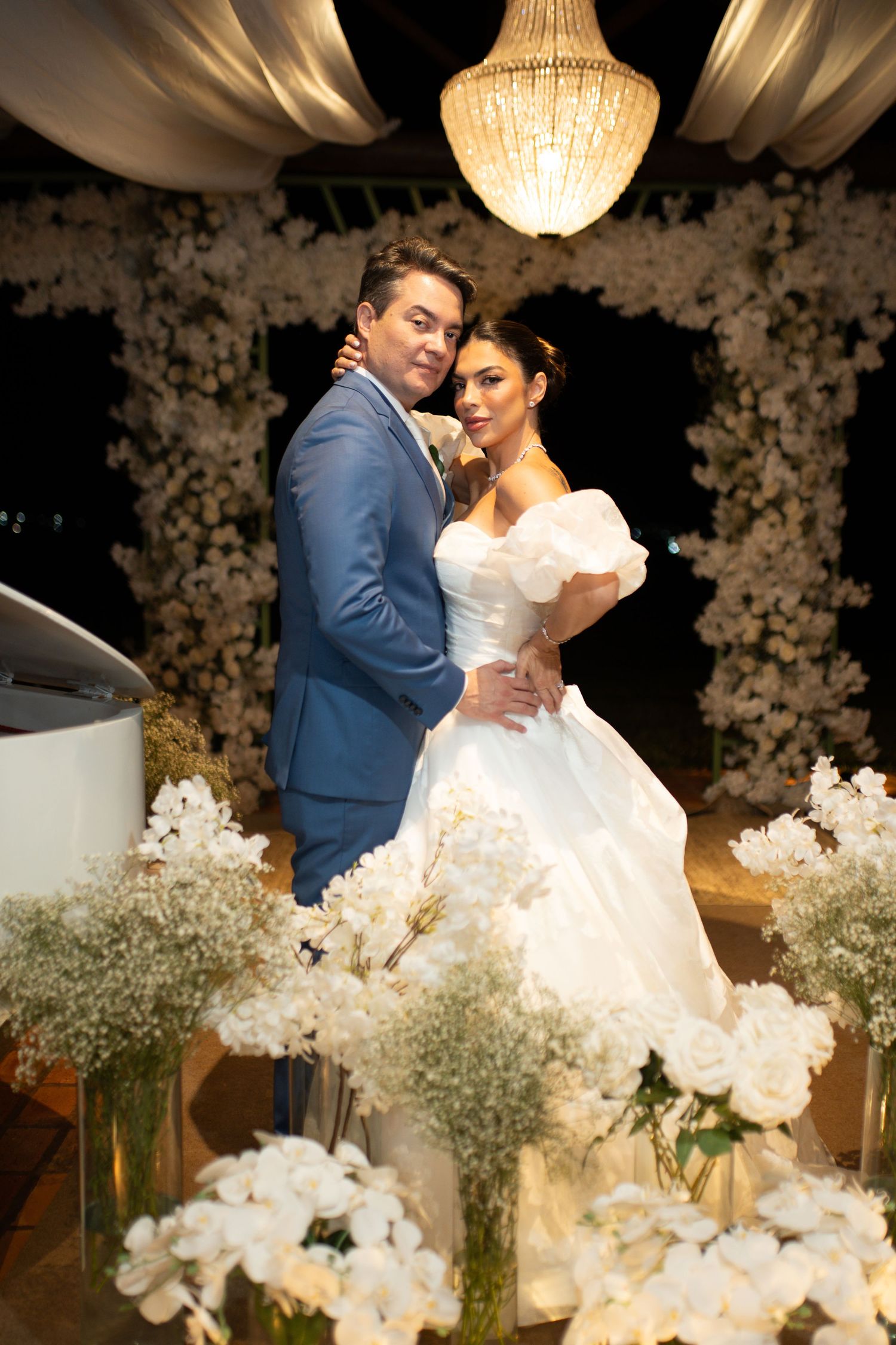 Elegant bride in white gown and groom in blue suit embracing at a floral wedding ceremony with chandelier backdrop.