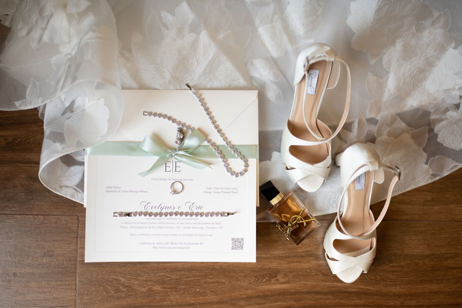 Bridal flat lay featuring white heels, pearl necklace, gold rings, perfume, and elegant wedding invitation on wood floor.