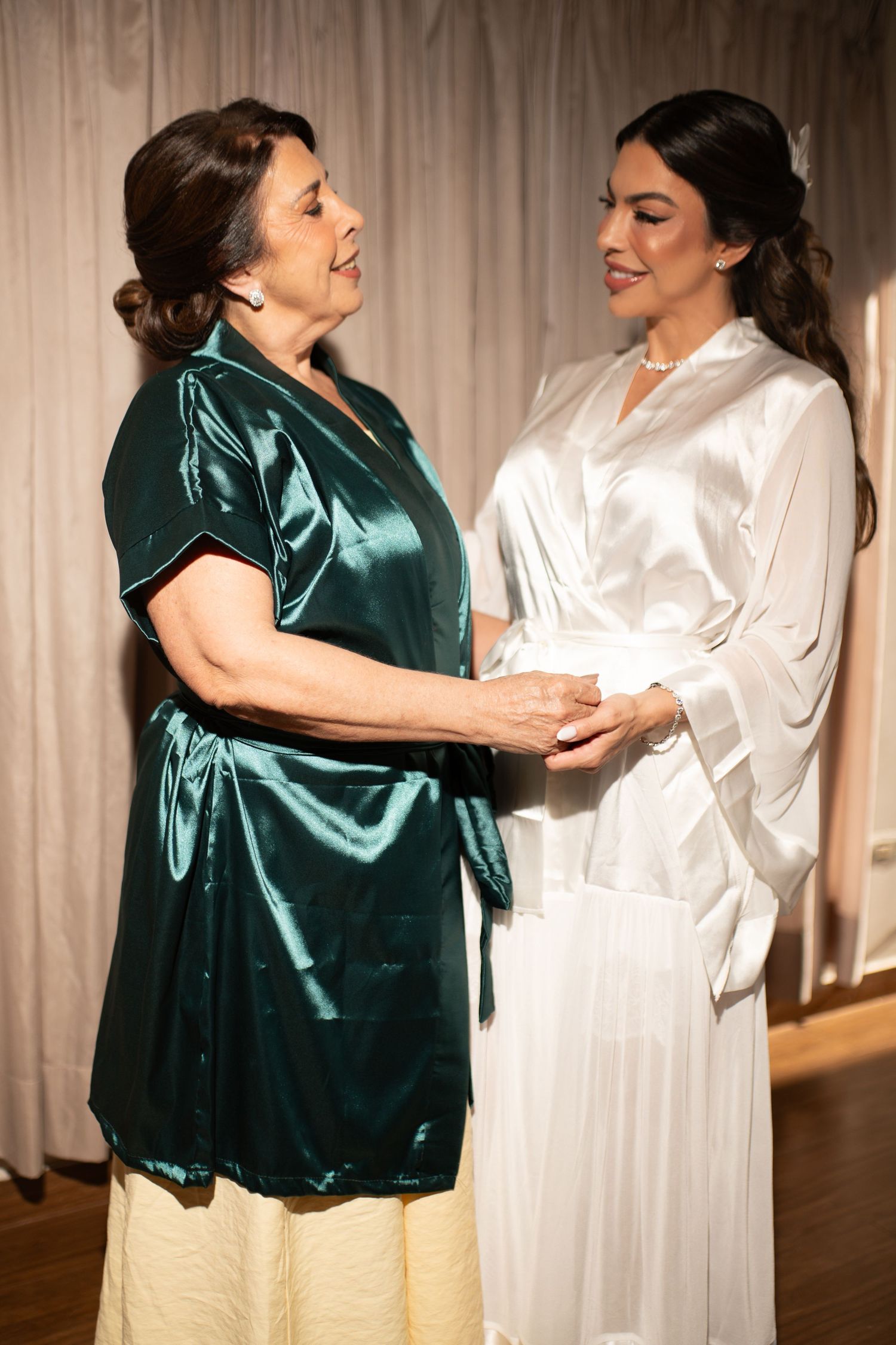Two women in satin robes holding hands, sharing a tender moment, possibly a bride and her mother before a wedding.