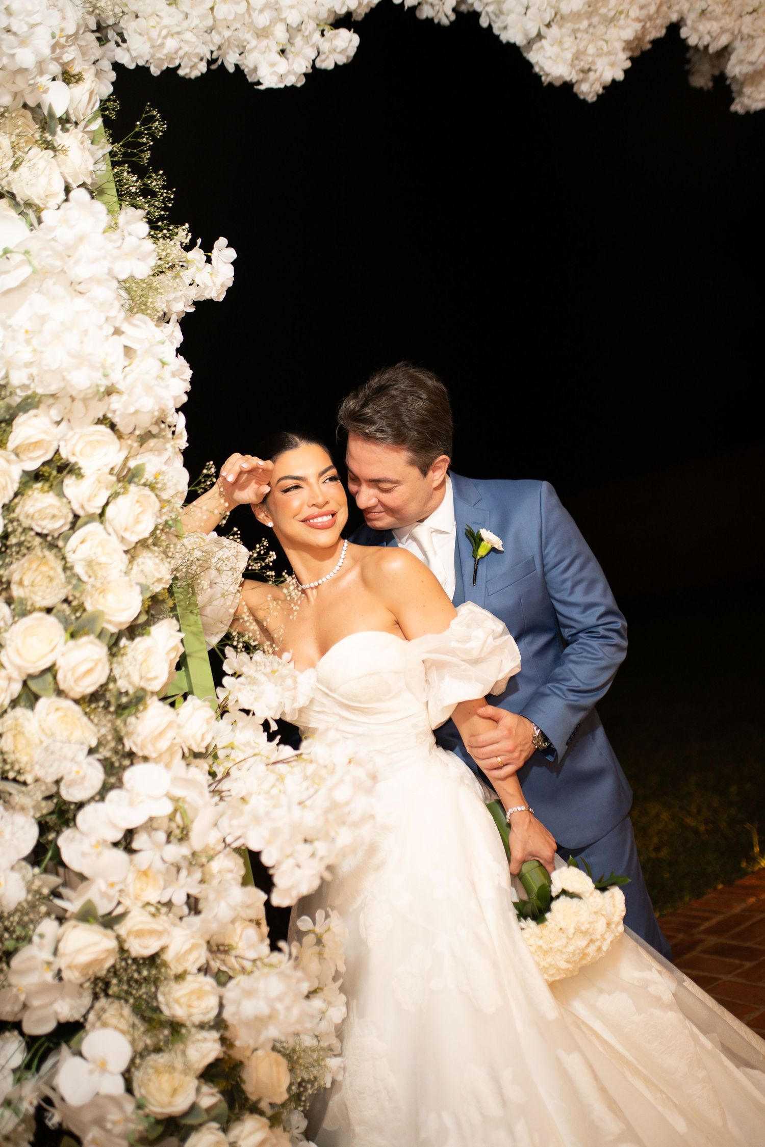 Bride and groom share a romantic moment under a white floral arch at an elegant nighttime wedding ceremony.