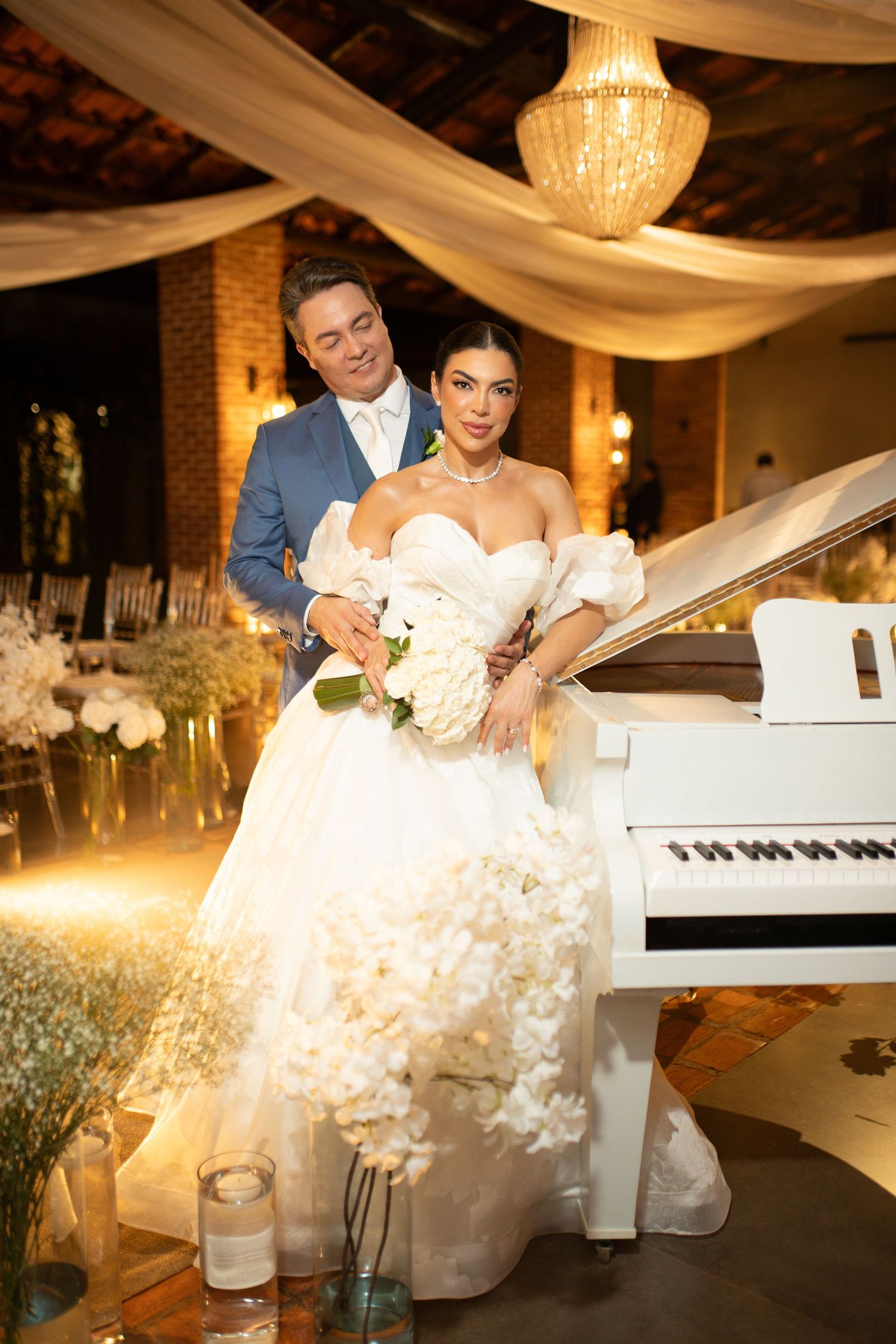 Elegant bride in white ballgown holding bouquet poses with groom in blue suit beside white grand piano at wedding reception.