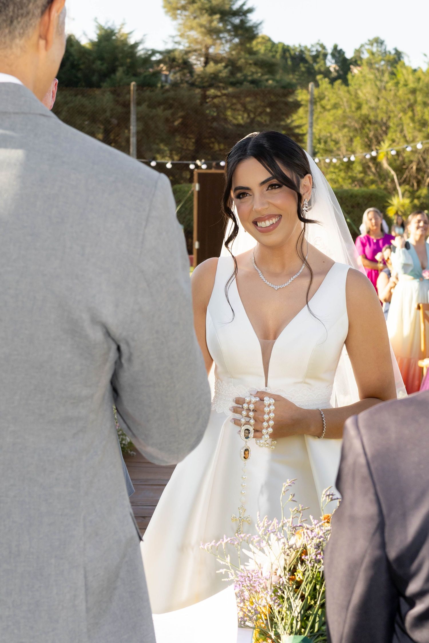 Smiling bride in white gown holding rosary beads at outdoor wedding ceremony with groom in gray suit.
