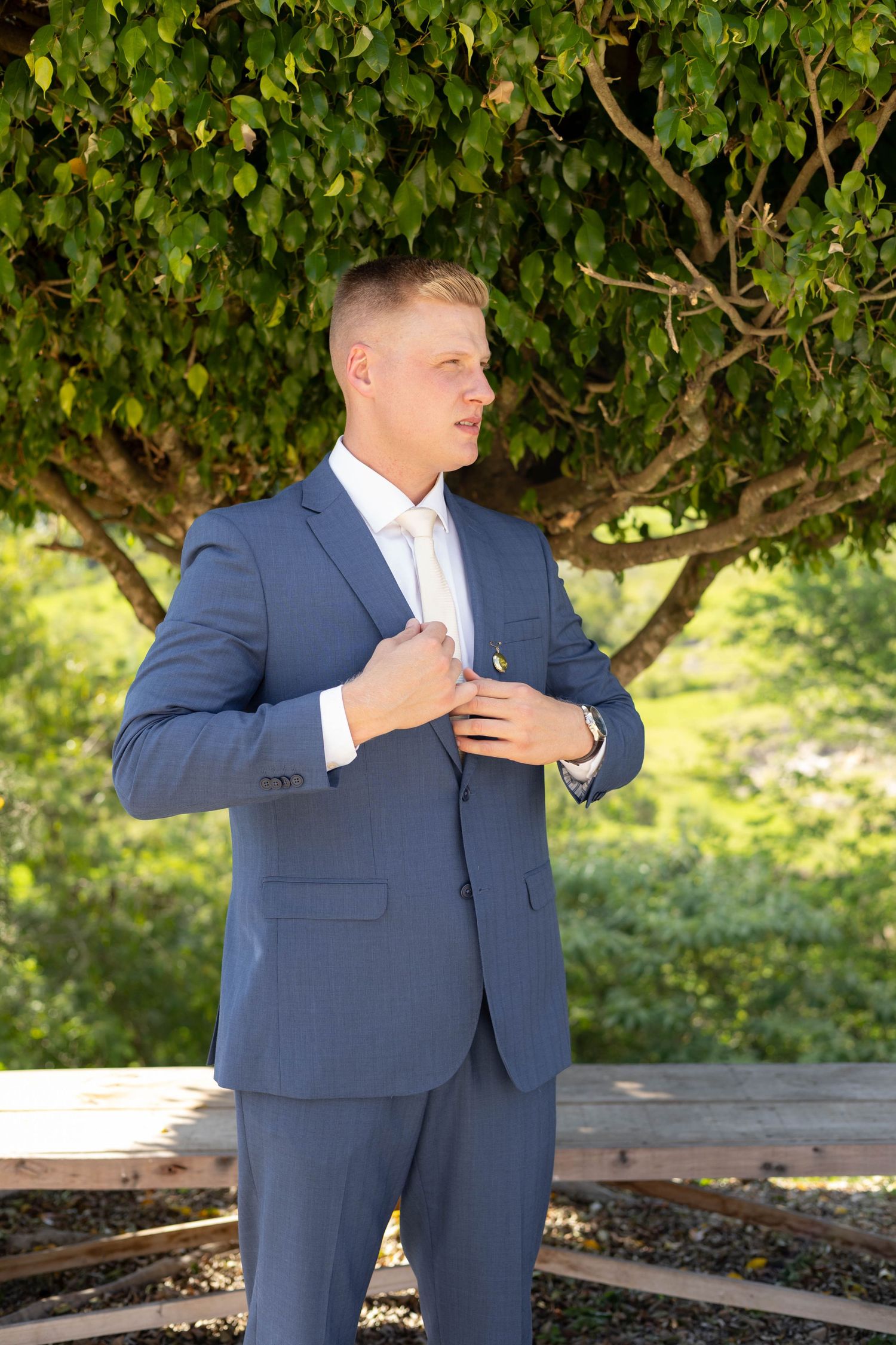 Man in blue suit adjusting jacket lapel outdoors with green tree background, likely a groom at a wedding.