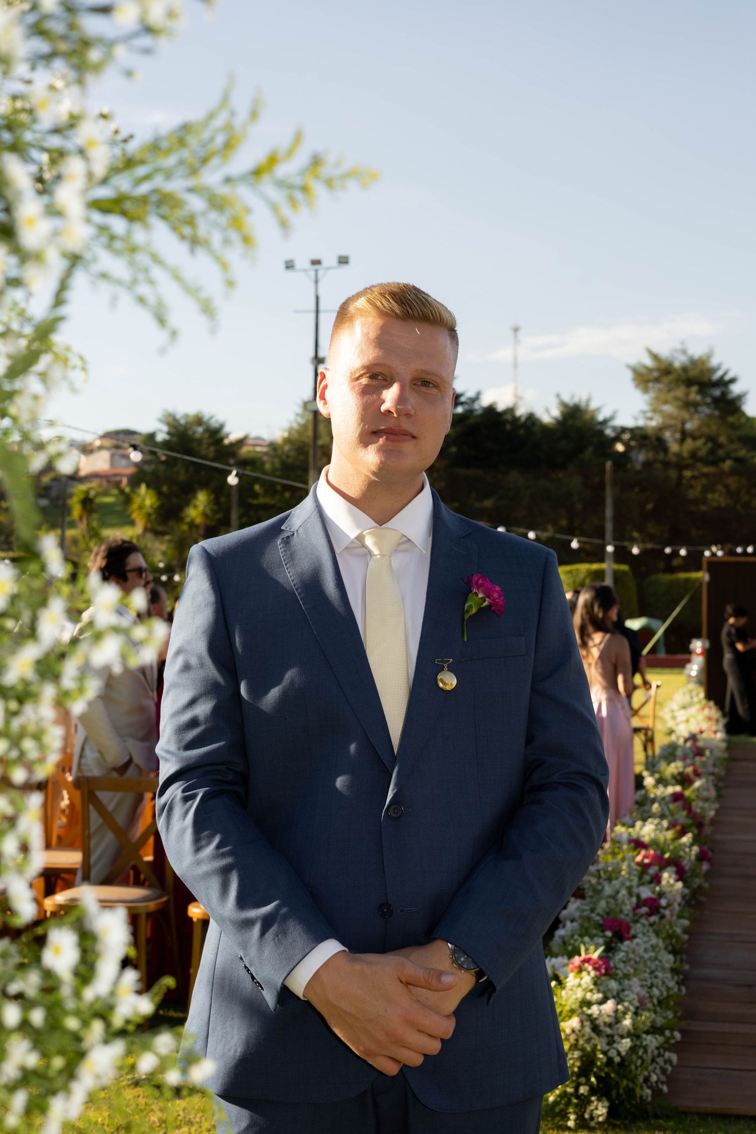 A groom in a navy blue suit with a pink boutonniere stands outdoors at a wedding ceremony on a sunny day.