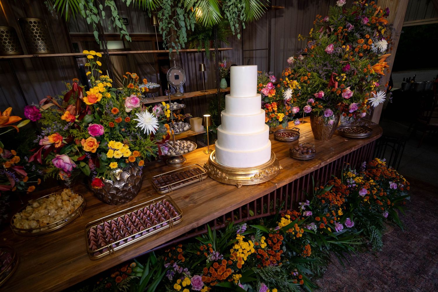 Elegant white tiered wedding cake on a wooden dessert table surrounded by vibrant colorful floral arrangements.