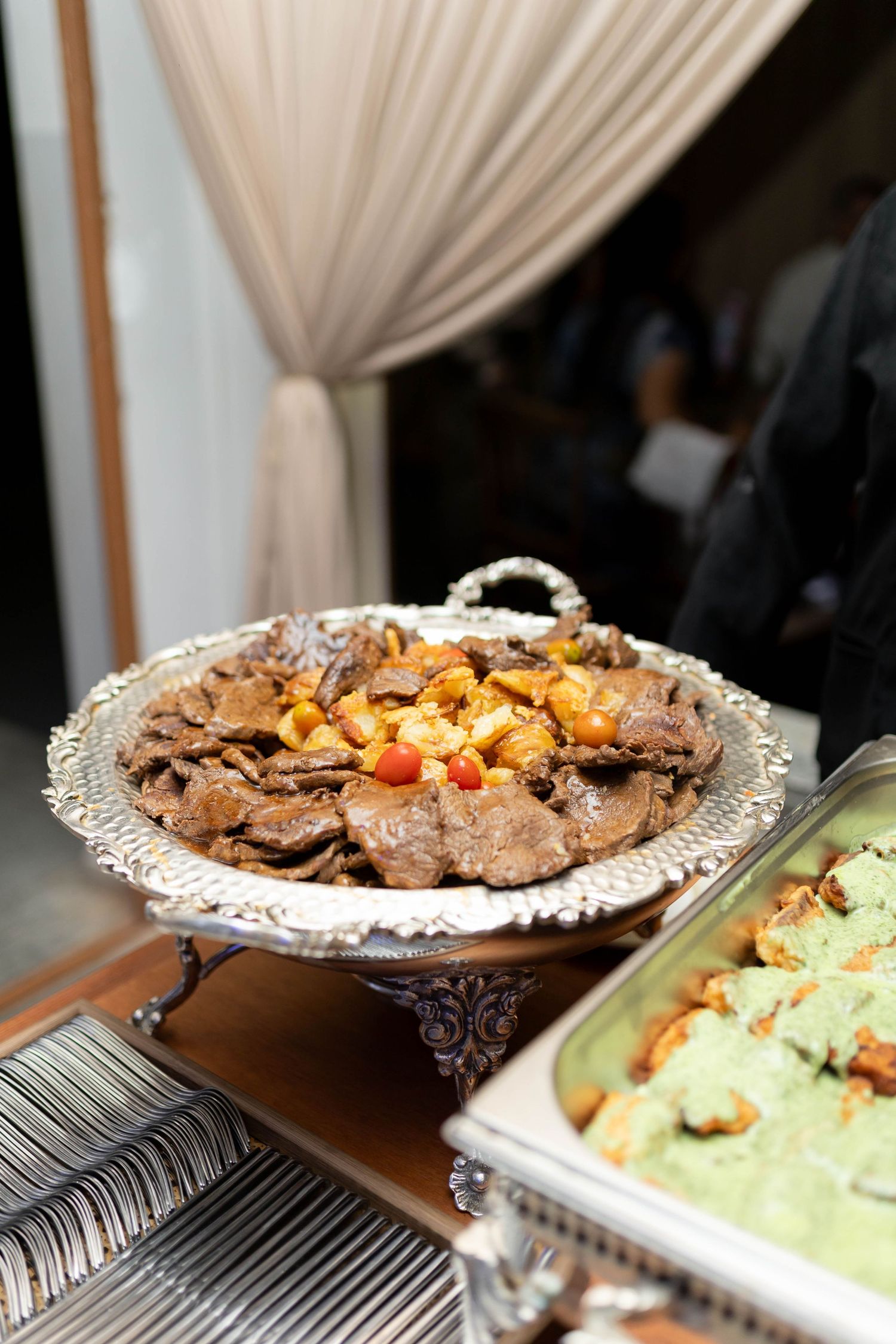Elegant silver platter with sliced meat and vegetables at a catered buffet event beside a green dish.