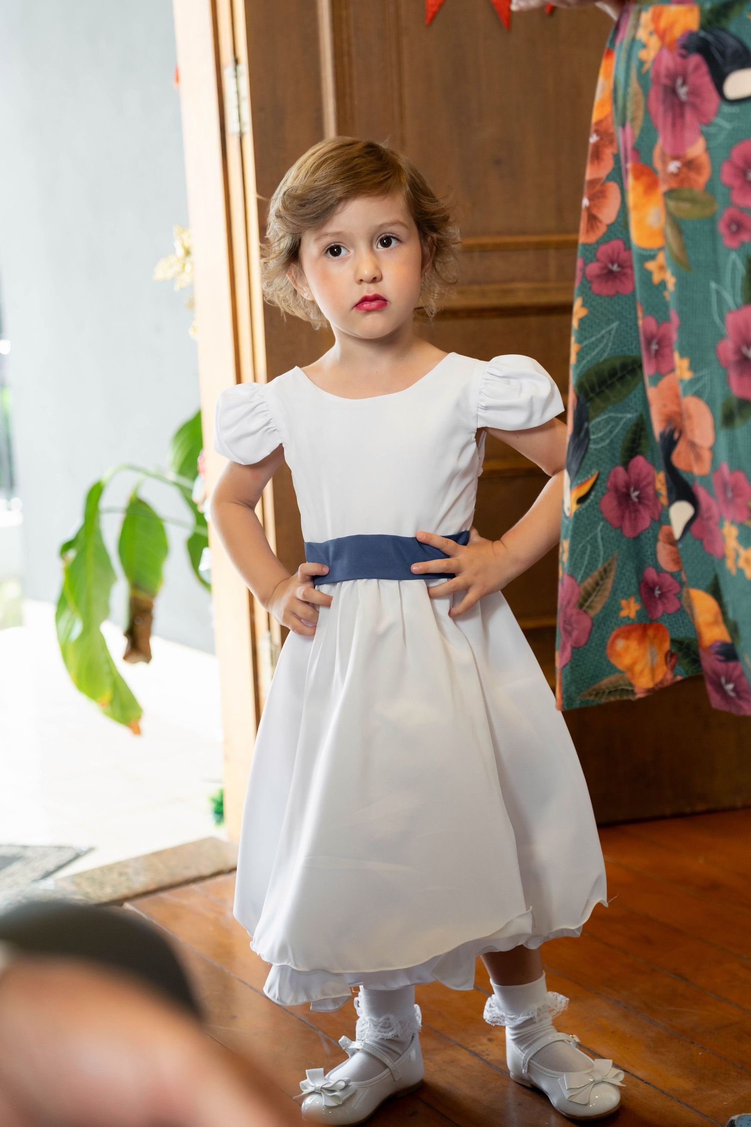 Little girl in white dress with blue sash stands with hands on hips in elegant room.