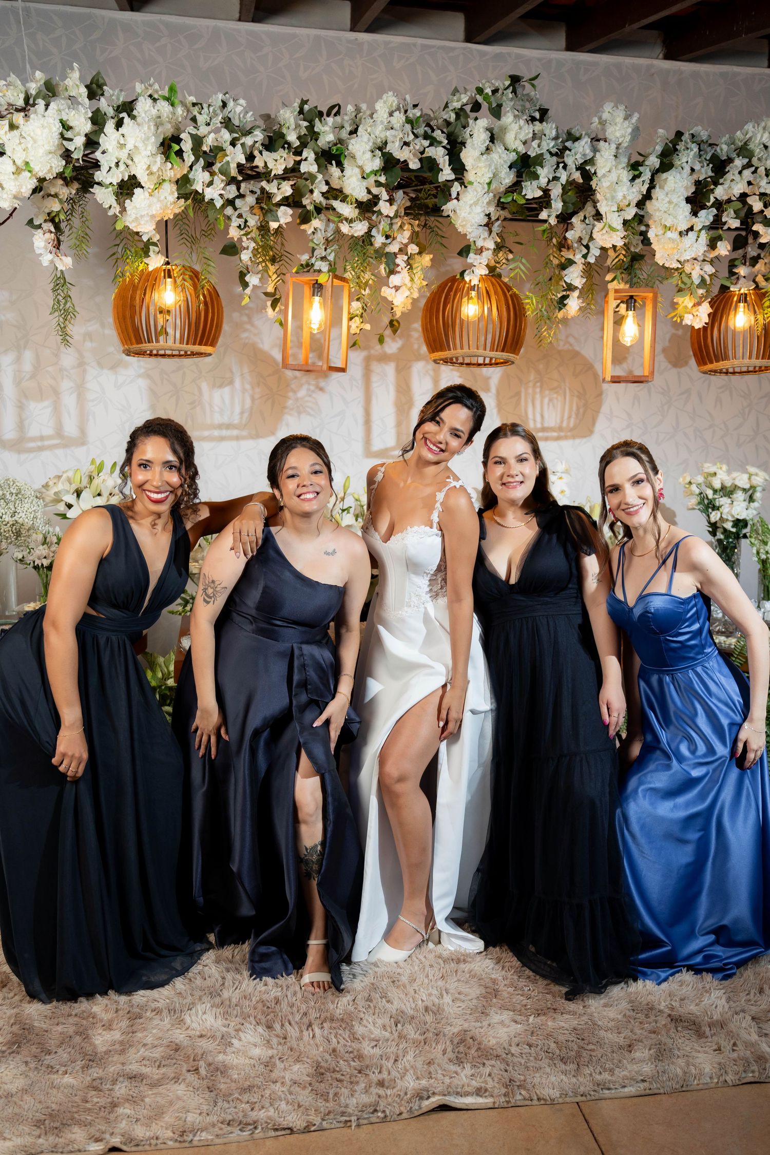 Bride in white gown poses with four bridesmaids in navy and blue dresses under floral decor and hanging lanterns.