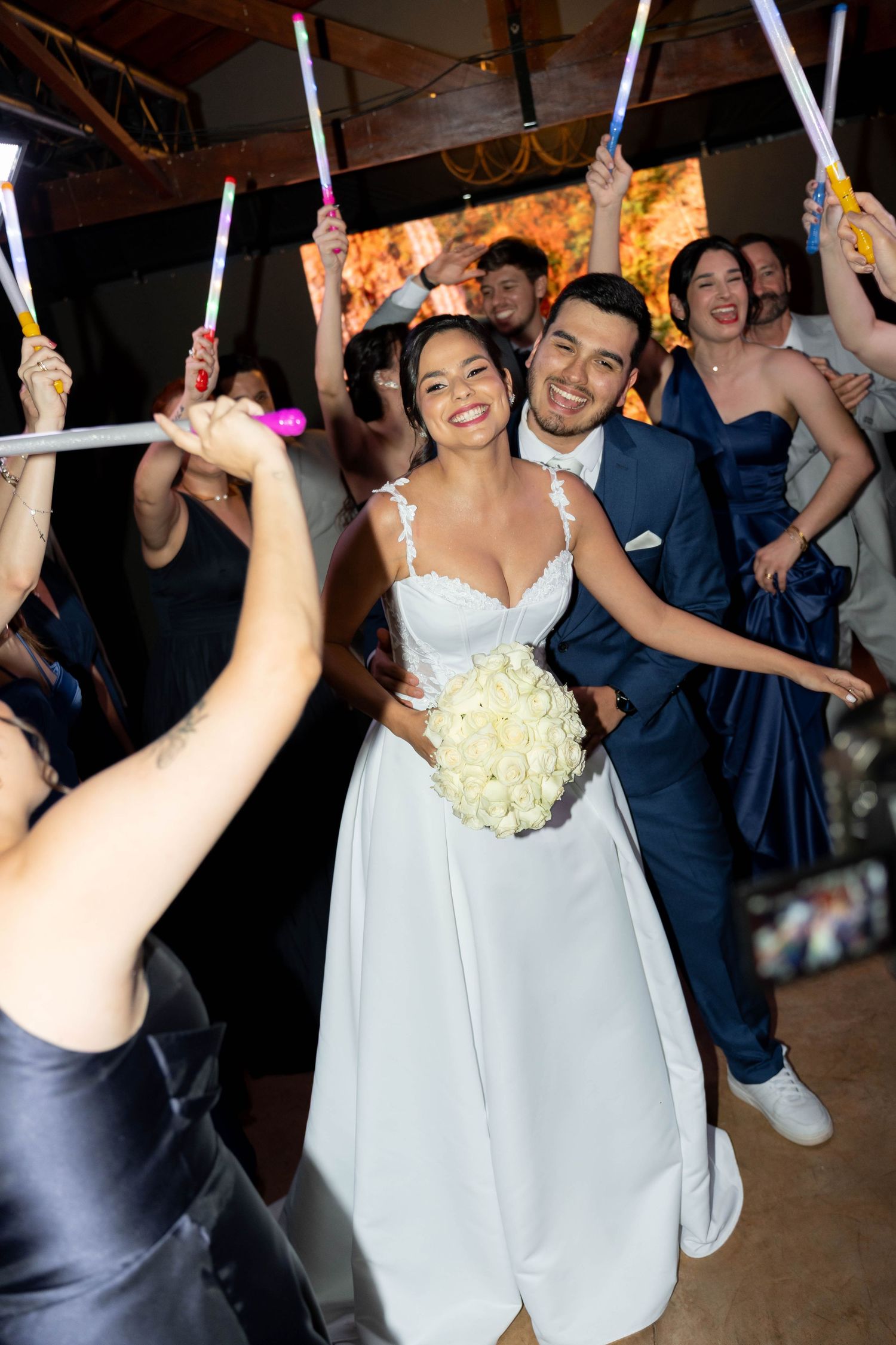 Bride and groom walk through a glowing wand send-off surrounded by cheering wedding guests indoors.