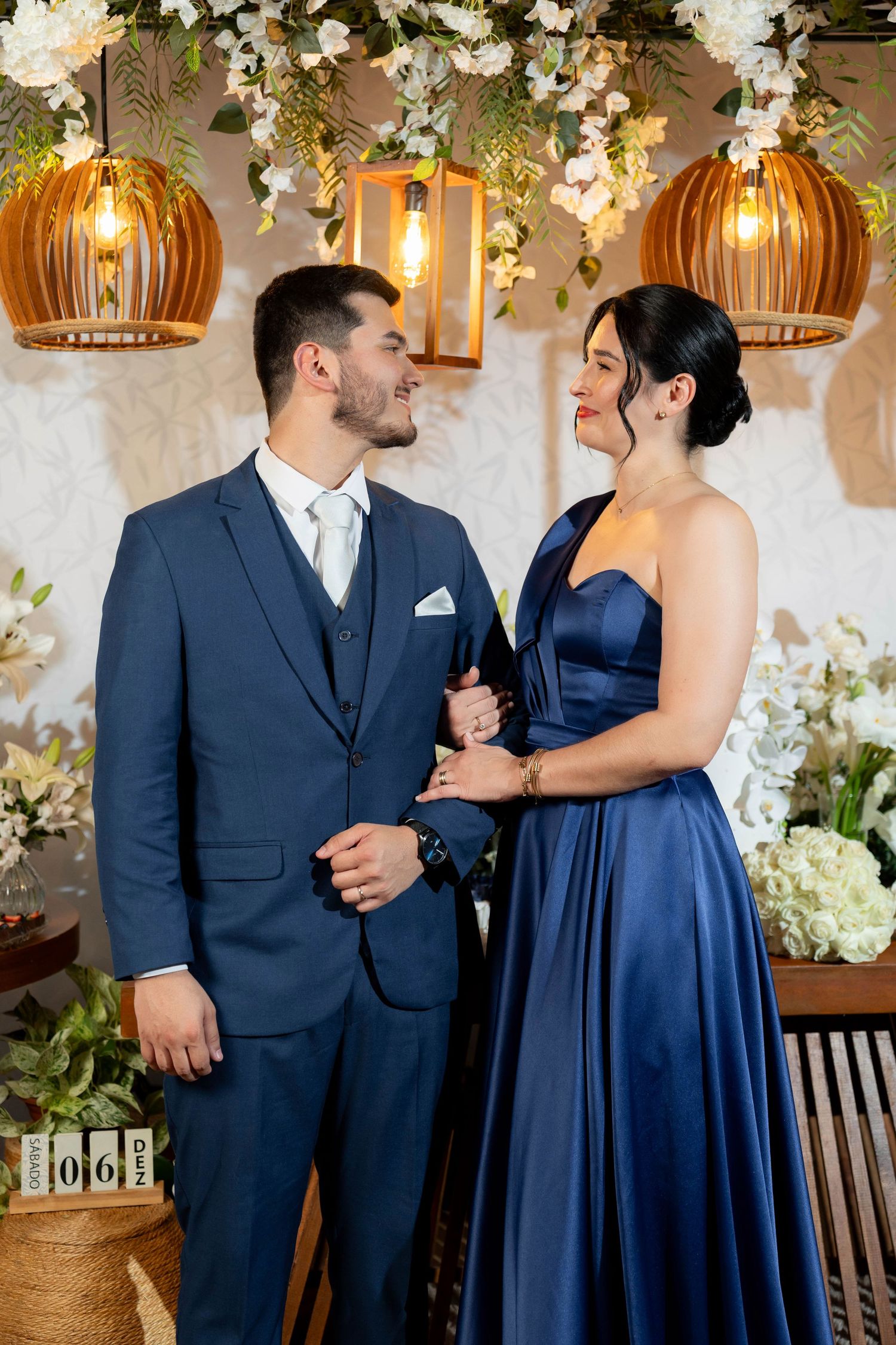 Elegant couple in matching navy blue outfits smiling at each other at a floral wedding venue with warm pendant lights.