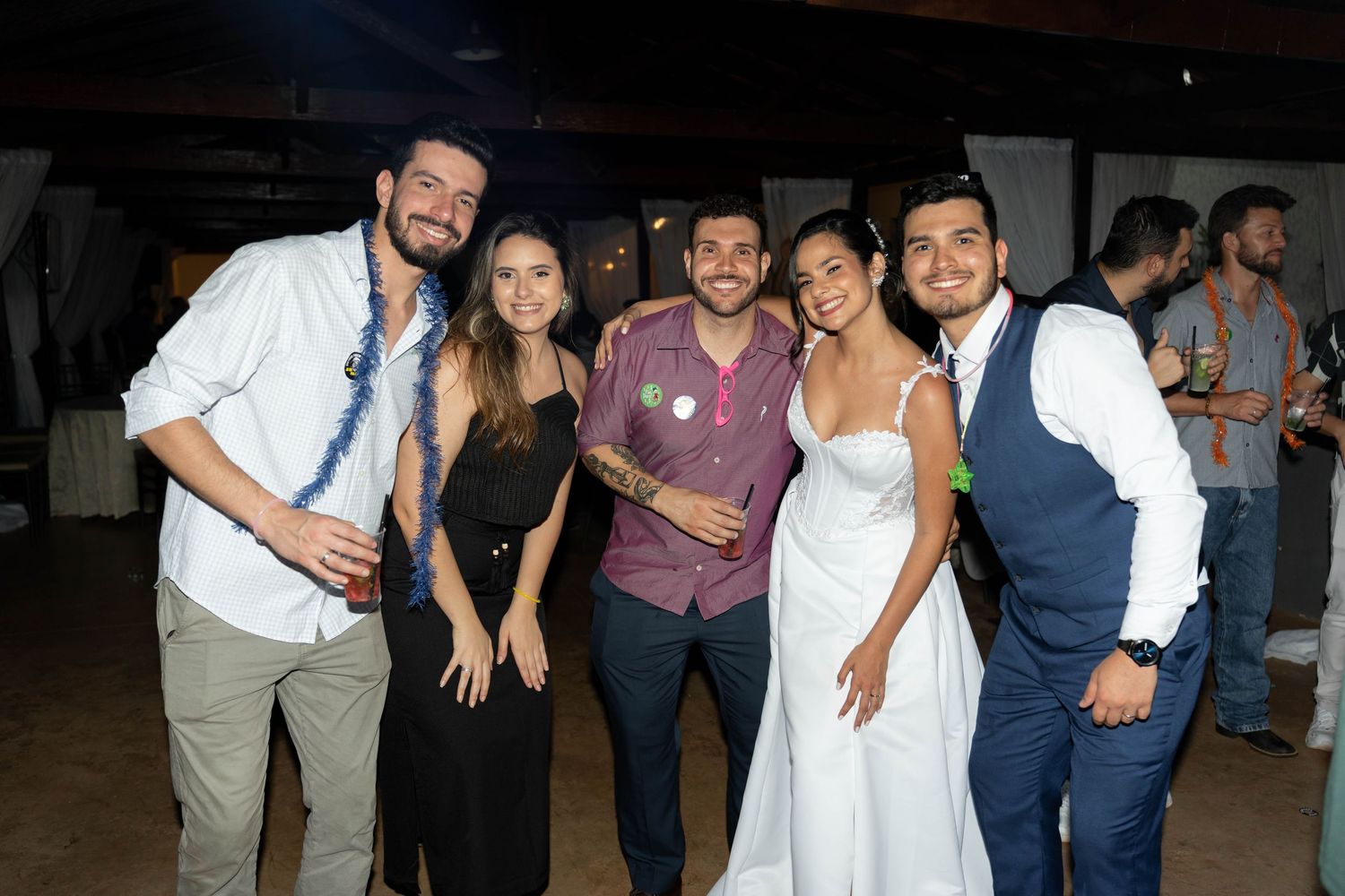 Five friends pose together smiling at a wedding reception, with the bride in a white dress standing in the center.