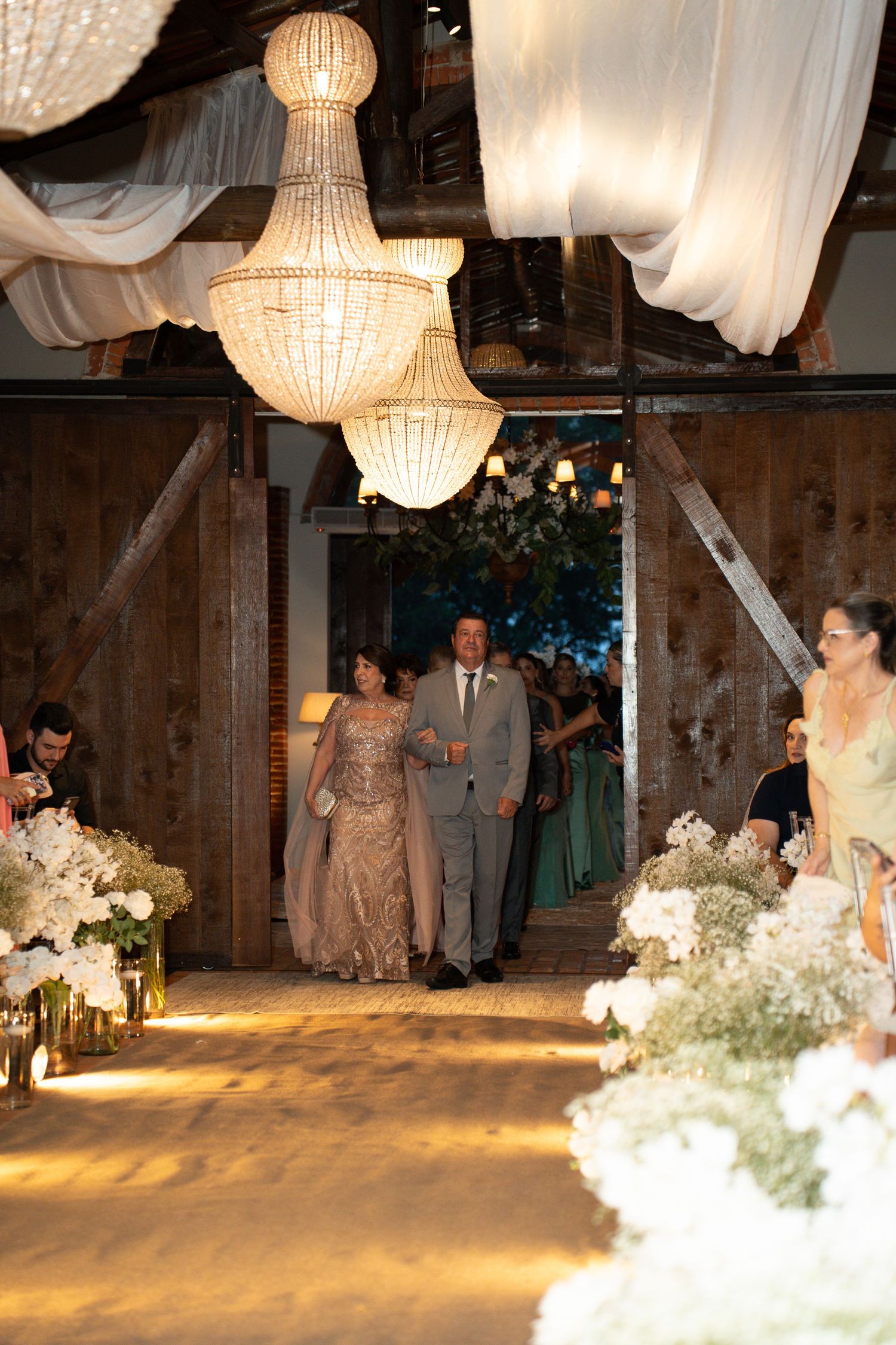 Elegant wedding ceremony entrance in a rustic barn with crystal chandeliers, floral arrangements, and guests walking down the aisle.