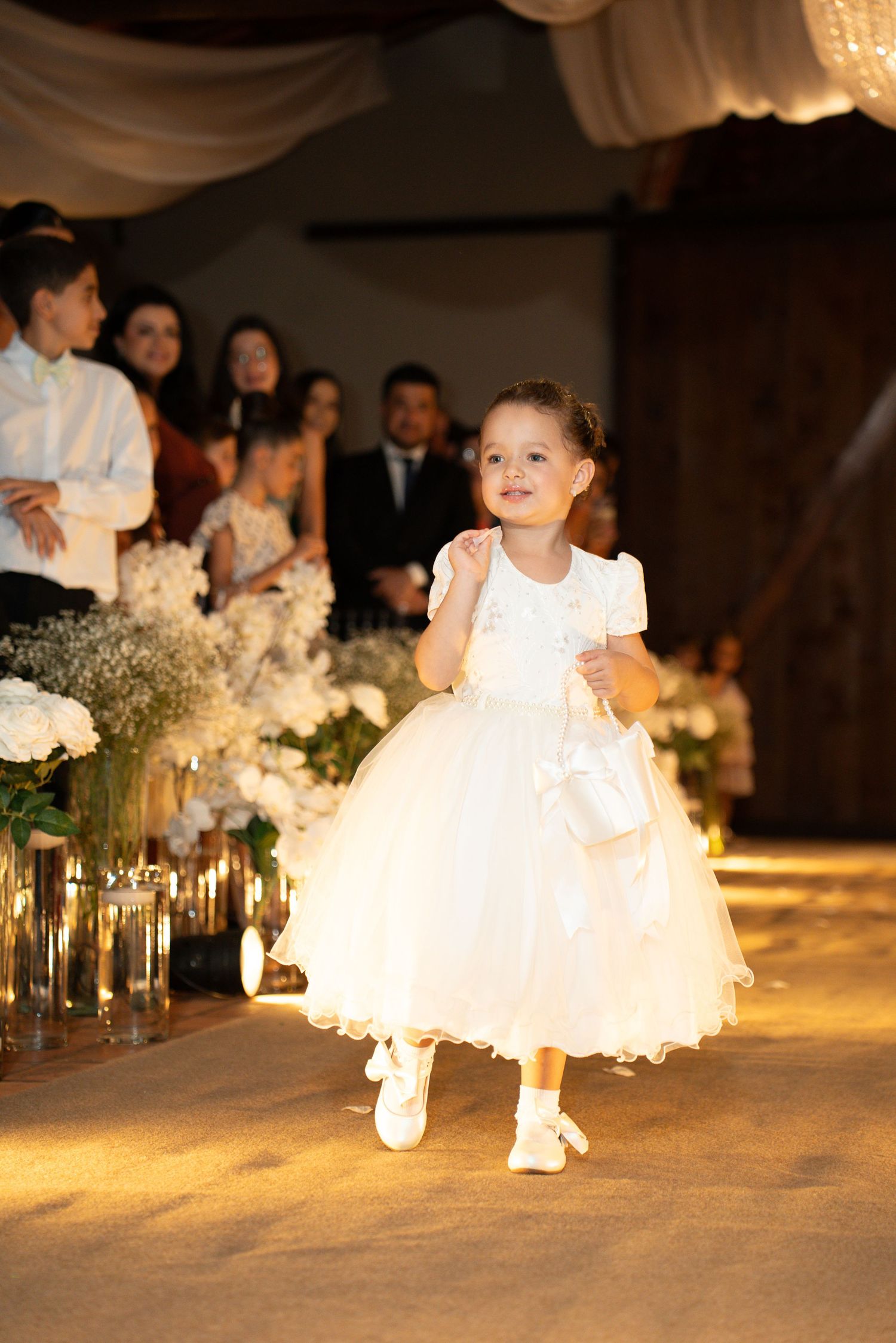 Adorable flower girl in white dress walking down wedding aisle, smiling joyfully at elegant indoor ceremony.