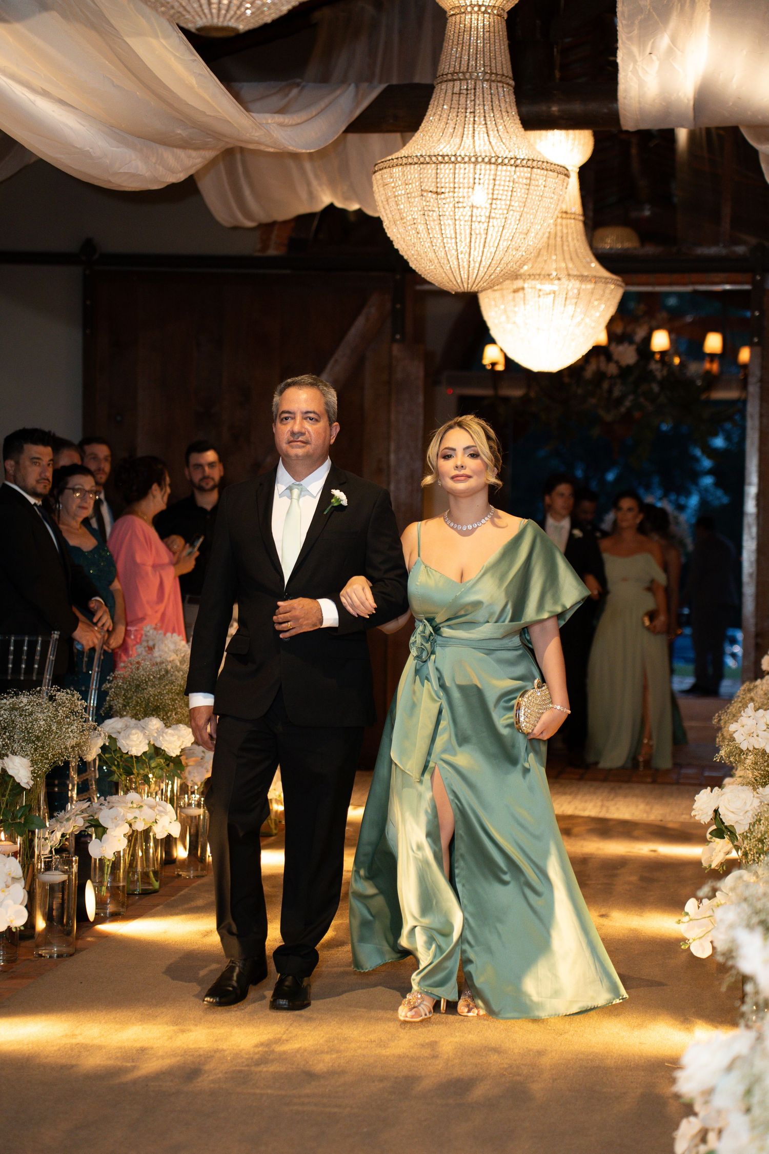 Elegant couple walking down wedding aisle, man in tuxedo and woman in teal satin gown, crystal chandeliers above.
