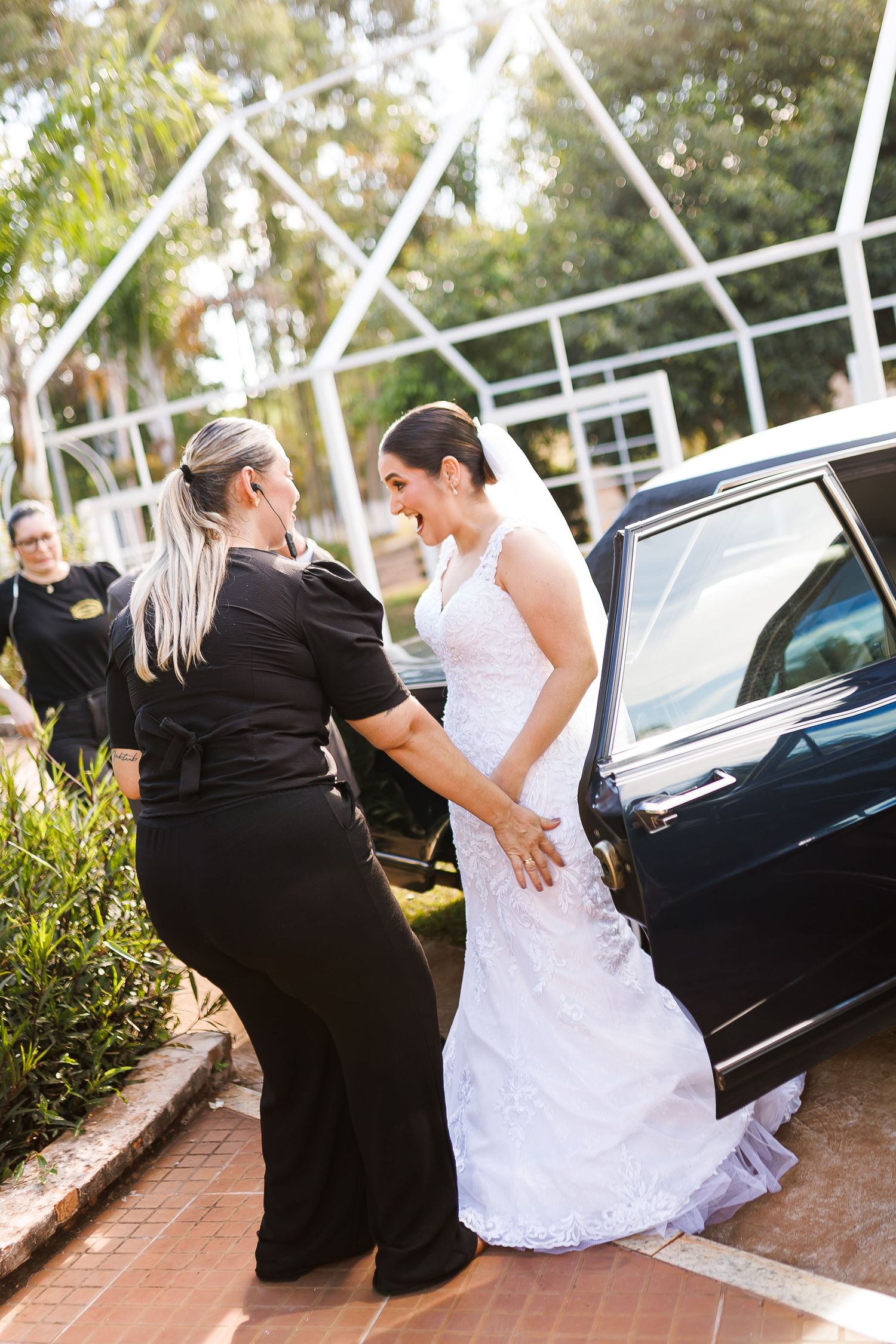 Bride in white lace gown assisted by woman in black near a dark car, with greenery and white structure in background.