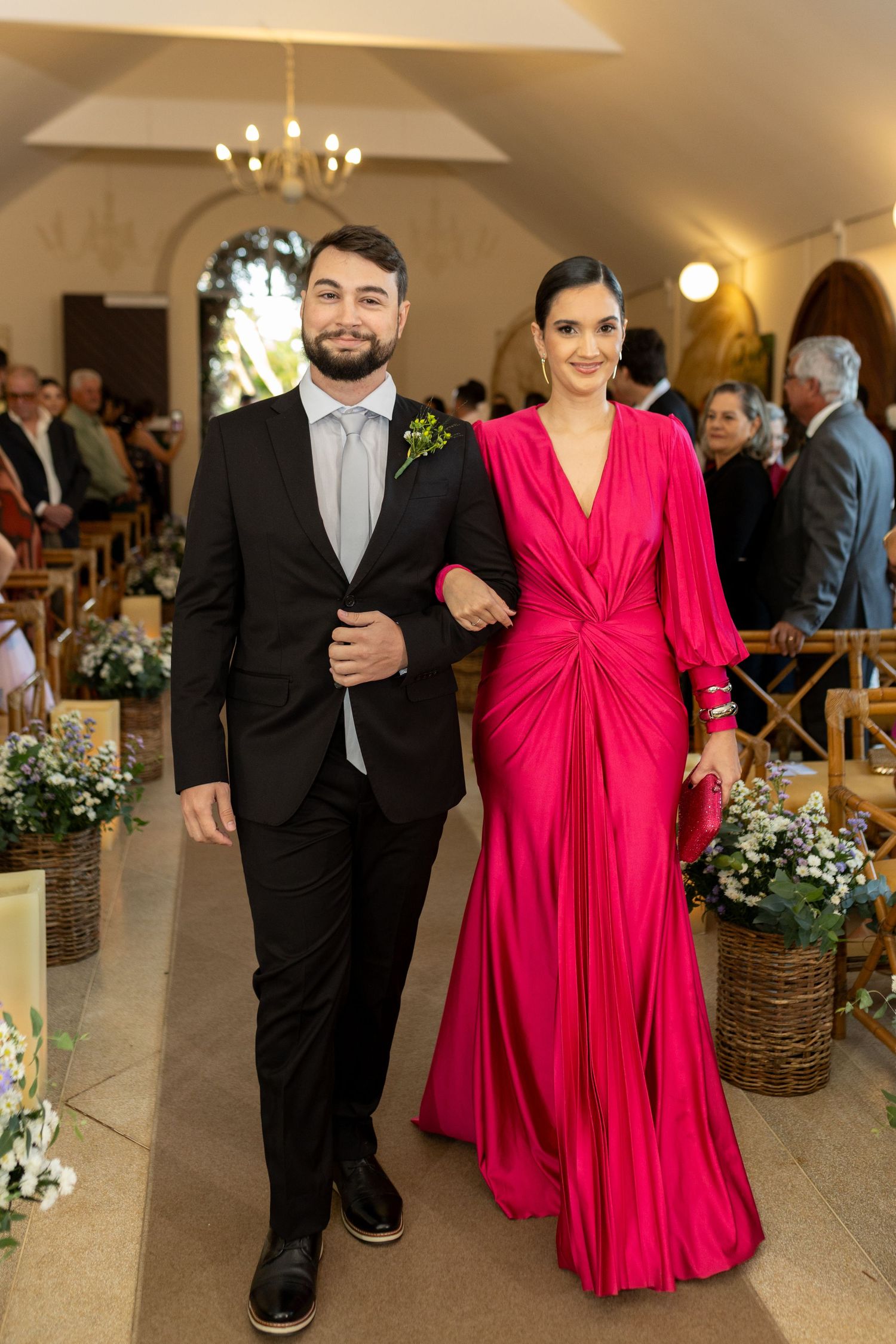 A man in a black suit and woman in a elegant red gown walk arm in arm down a church aisle.