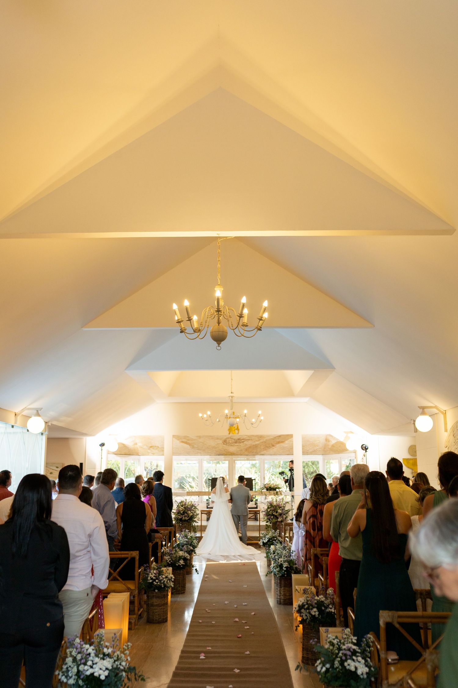 Elegant wedding ceremony in a white chapel with vaulted ceiling, chandelier, floral aisle decor, and guests standing as bride walks down.