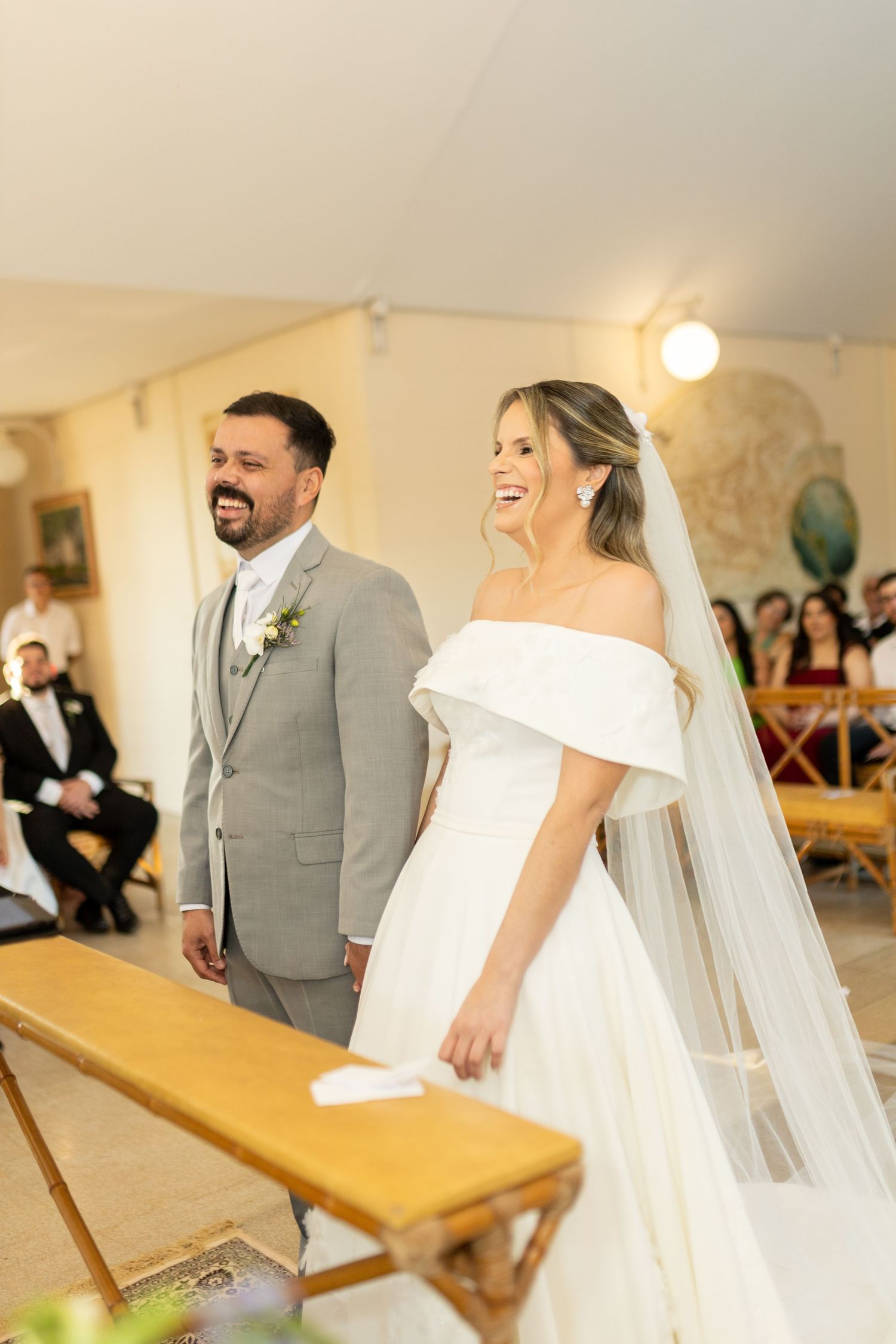 Smiling bride in off-shoulder white gown and veil stands beside groom in gray suit during wedding ceremony.