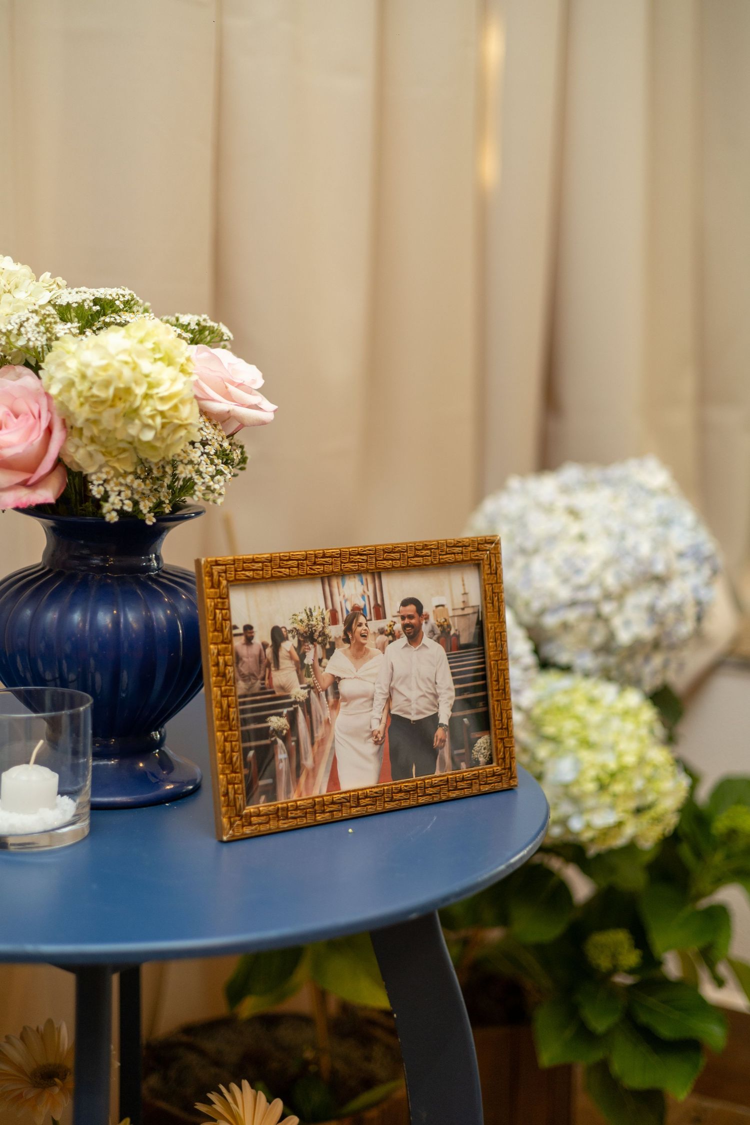 Blue table with framed wedding photo, blue vase of pink roses, white hydrangeas, and a candle in elegant decor.