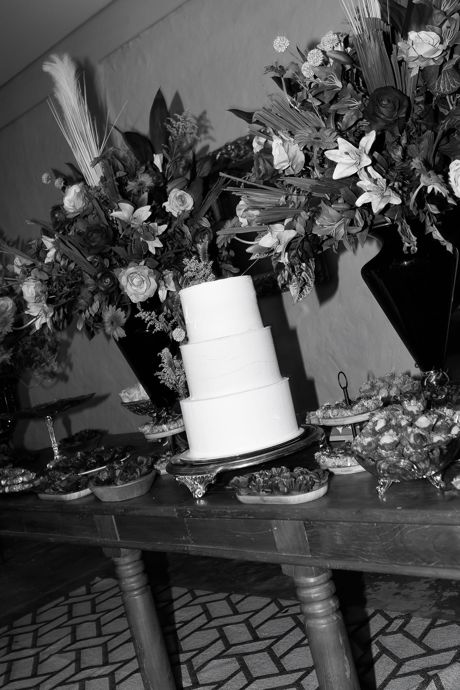 Black and white photo of a three-tier wedding cake on a wooden table surrounded by floral arrangements and desserts.