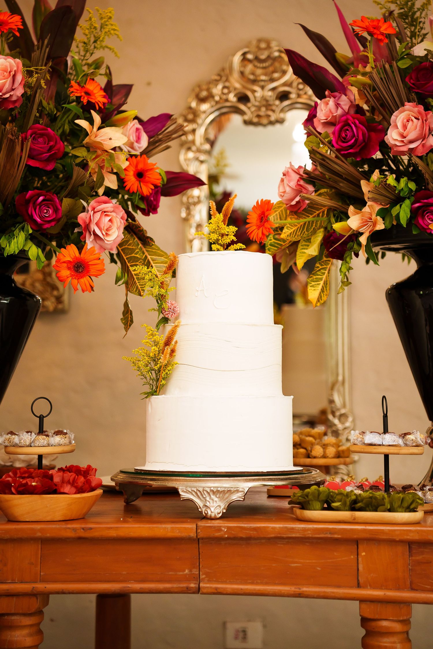 Elegant three-tier white wedding cake on wooden table with colorful floral arrangements and desserts.