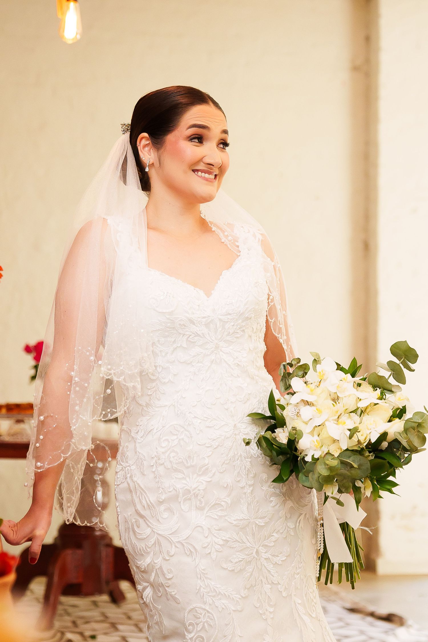 Smiling bride in white lace gown and veil holding white floral bouquet on her wedding day.