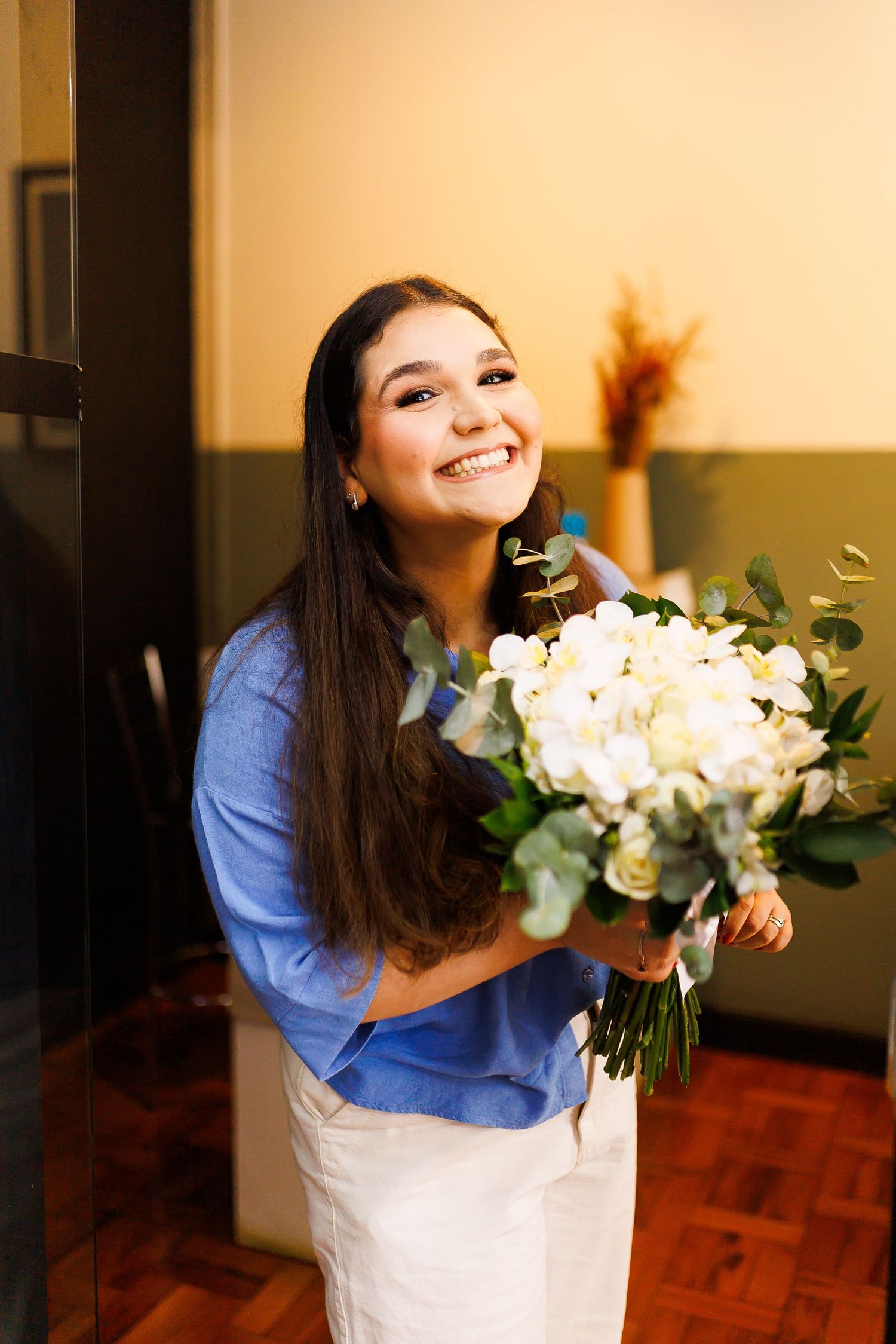 Smiling young woman holding a bouquet of white roses and greenery, wearing a blue shirt indoors.