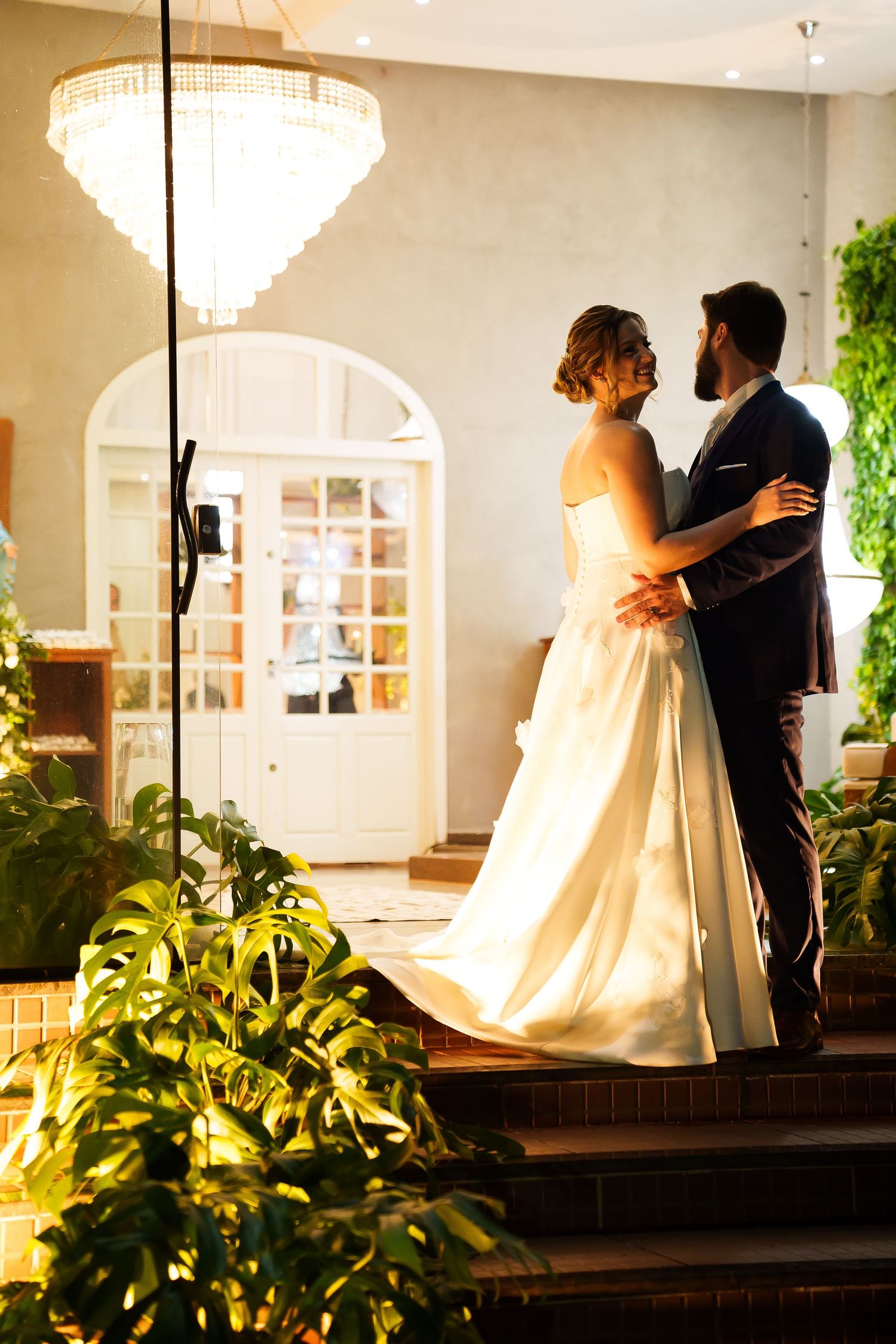 Bride and groom share a romantic embrace in elegant venue with chandelier and lush greenery.