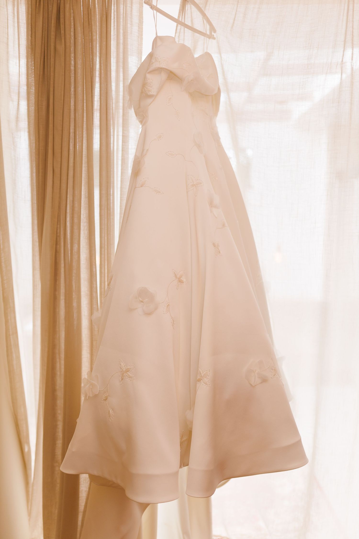 Elegant white strapless A-line wedding dress with floral appliques hanging on a hanger against sheer curtains.