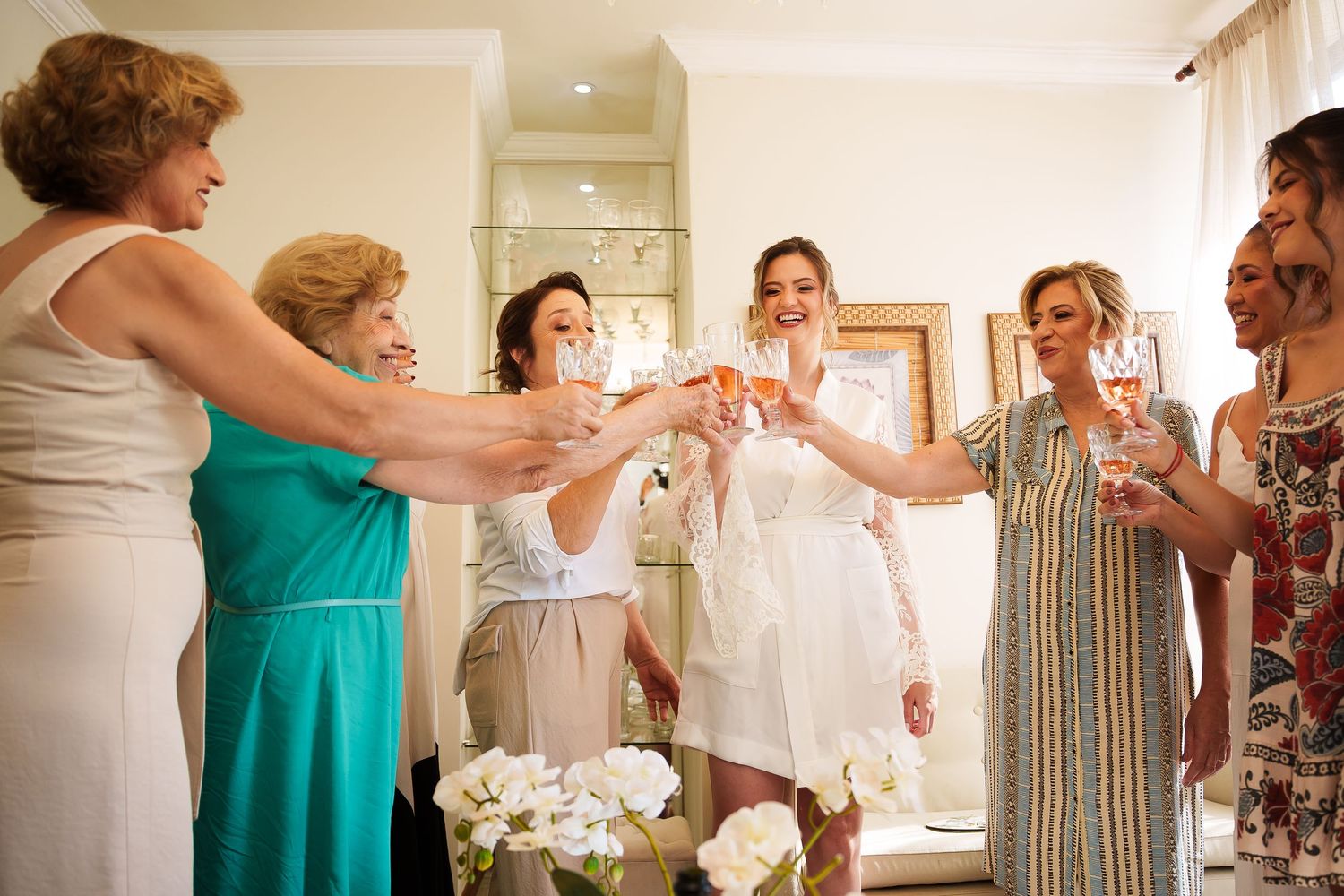 Bride in white robe toasting champagne with bridesmaids and family members during wedding preparations.