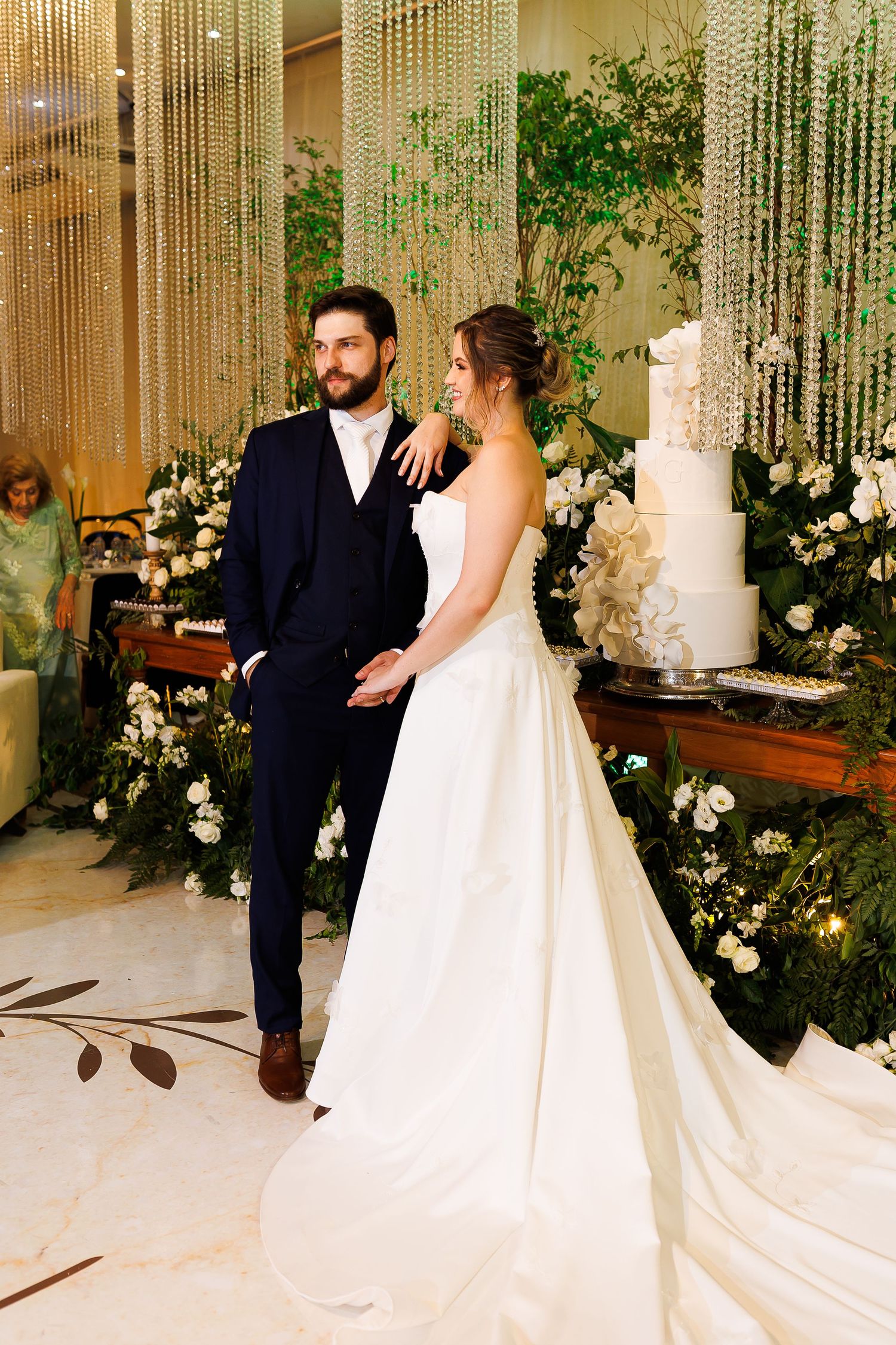 Elegant bride and groom posing together at wedding reception with white floral arrangements and tiered cake backdrop.