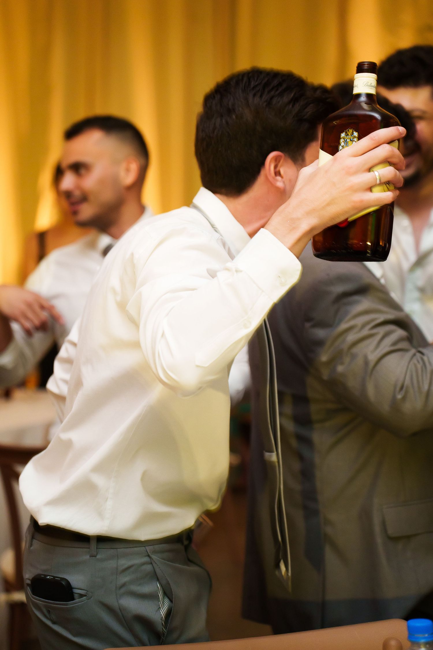 Man in white shirt drinking from a large bottle of whiskey at a party with friends in background.