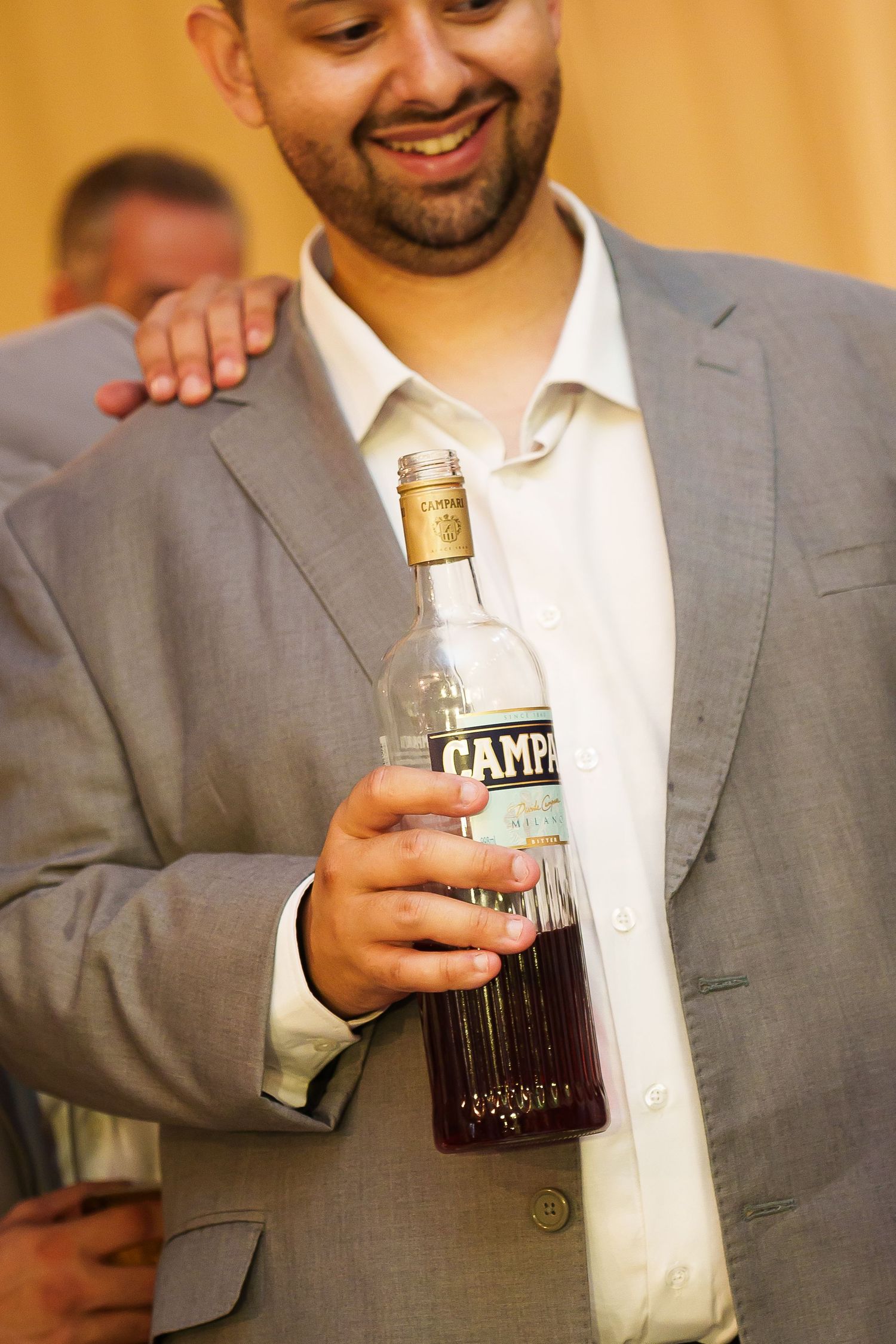 Smiling man in gray suit holding a bottle of Campari at a social event.