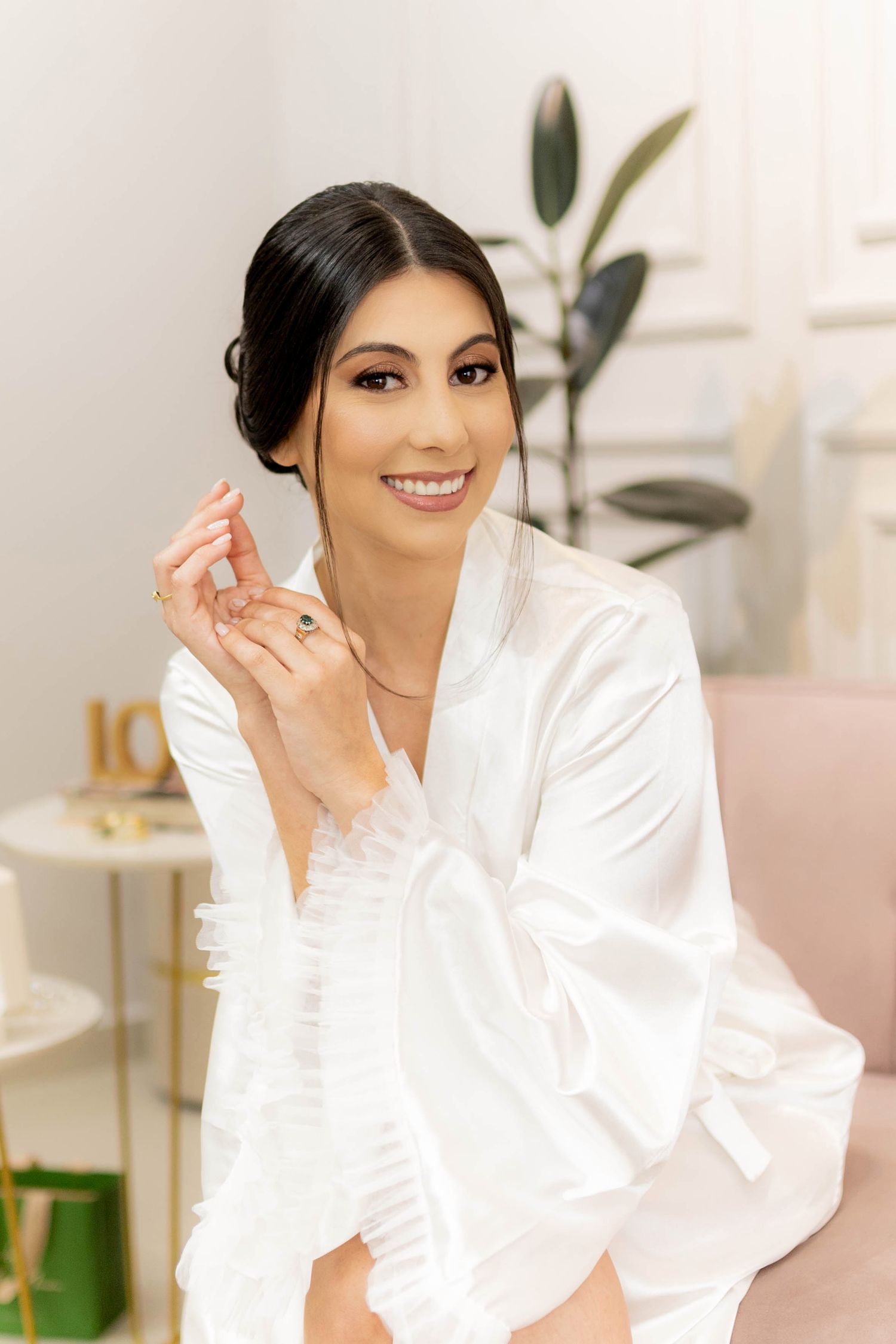 Smiling bride in white satin robe showing engagement ring while getting ready in a bright, elegant room.