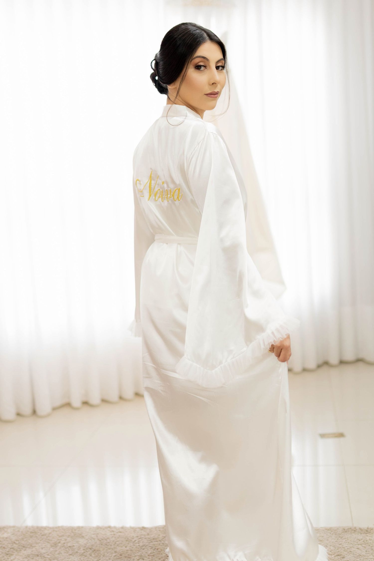 Bride in white satin robe with gold personalized name script and lace trim, wearing veil, posing elegantly indoors.