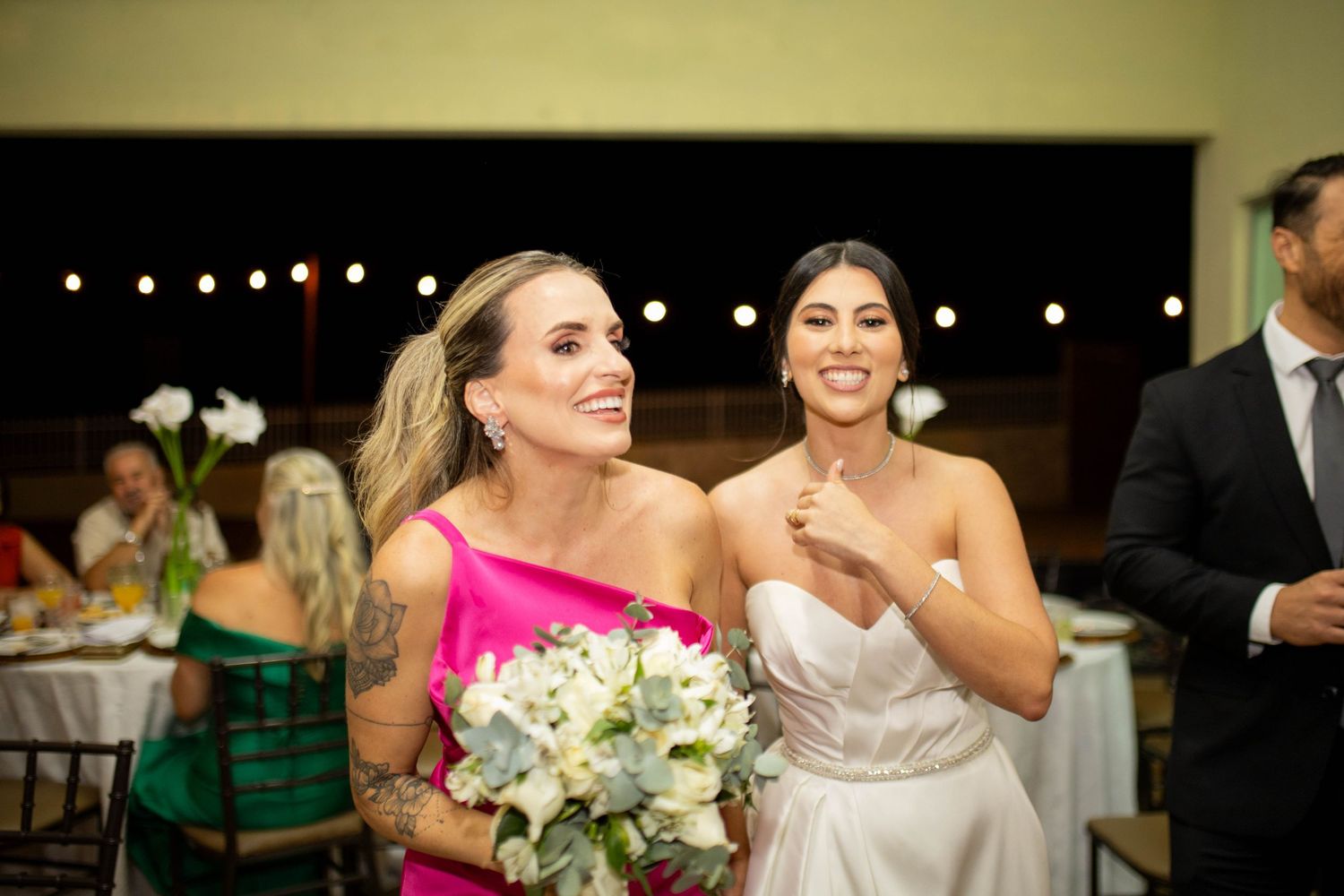 Two women smiling at a wedding reception, one in pink holding white flowers, the other in a white bridal gown.