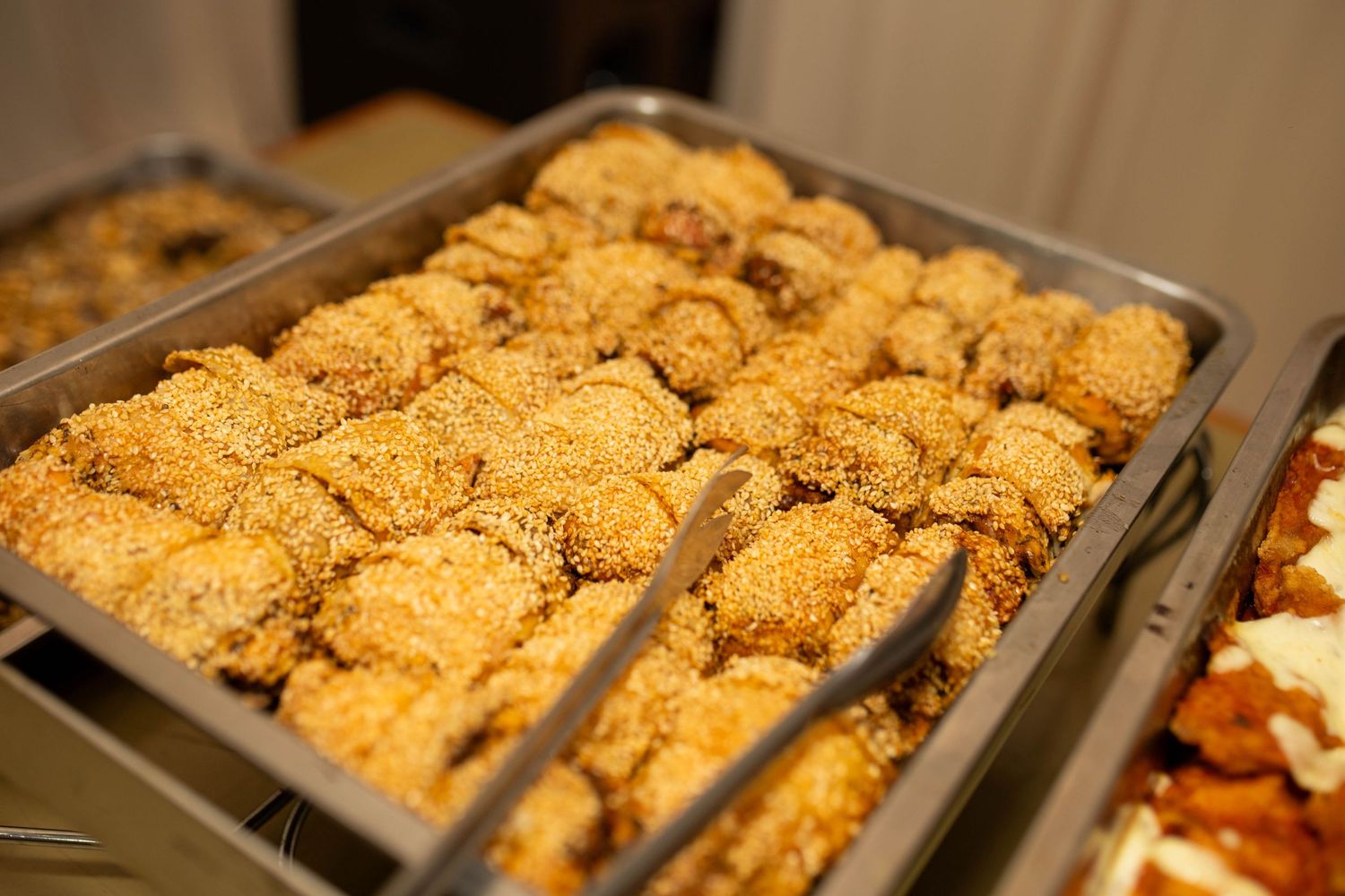 Sesame-topped baked pastries arranged in a metal catering tray at a buffet event.